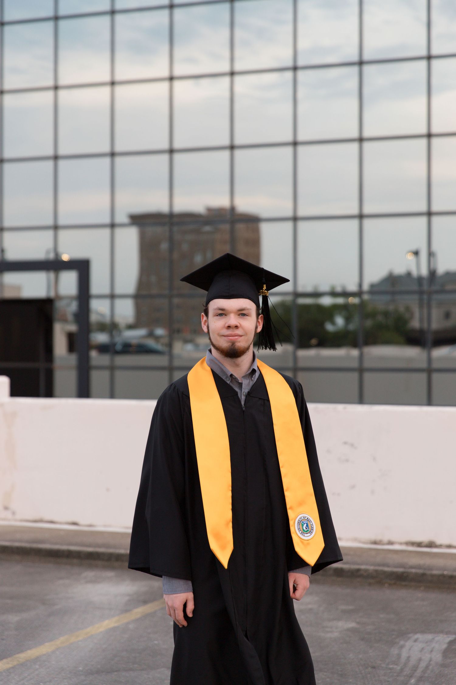 Graduate wearing black cap and gown with gold stole standing against modern architecture.