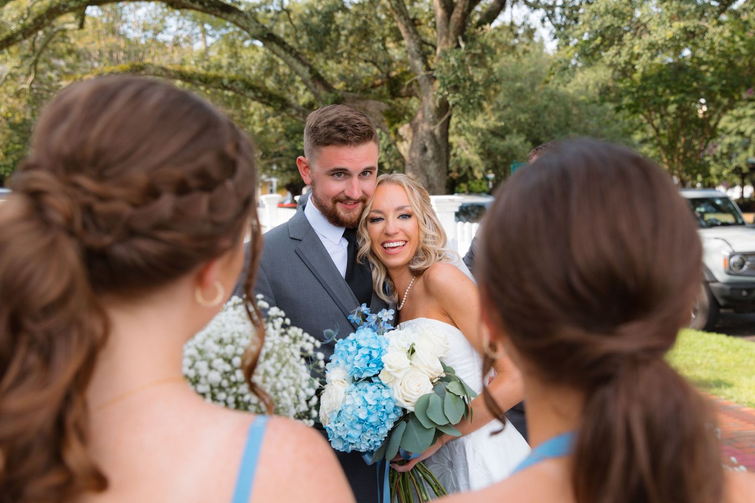 Newlyweds share a kiss while bridesmaids in blue dresses look on, holding blue and white bouquets.