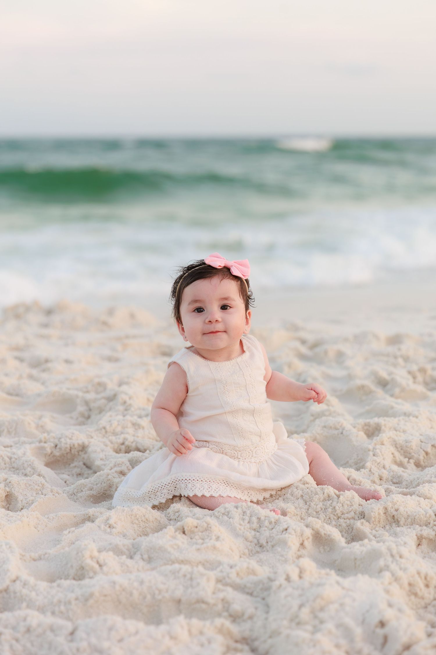 Baby girl in white dress and pink bow sits on sandy beach with ocean waves in background.