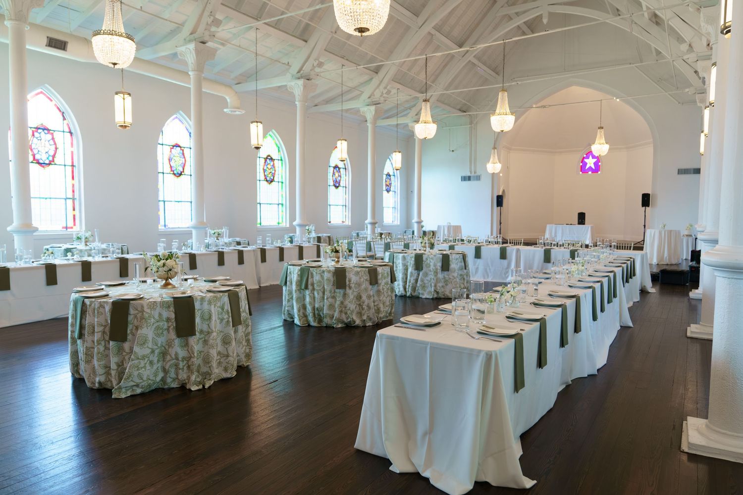 Elegant wedding reception hall with white tablecloths, green runners, arched windows, and pendant lights in a converted church venue.