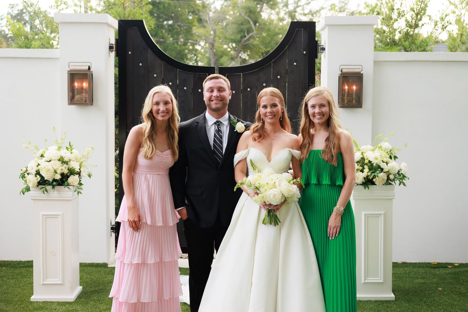 Bride in white gown poses with groom and two women in pink and green dresses outdoors.