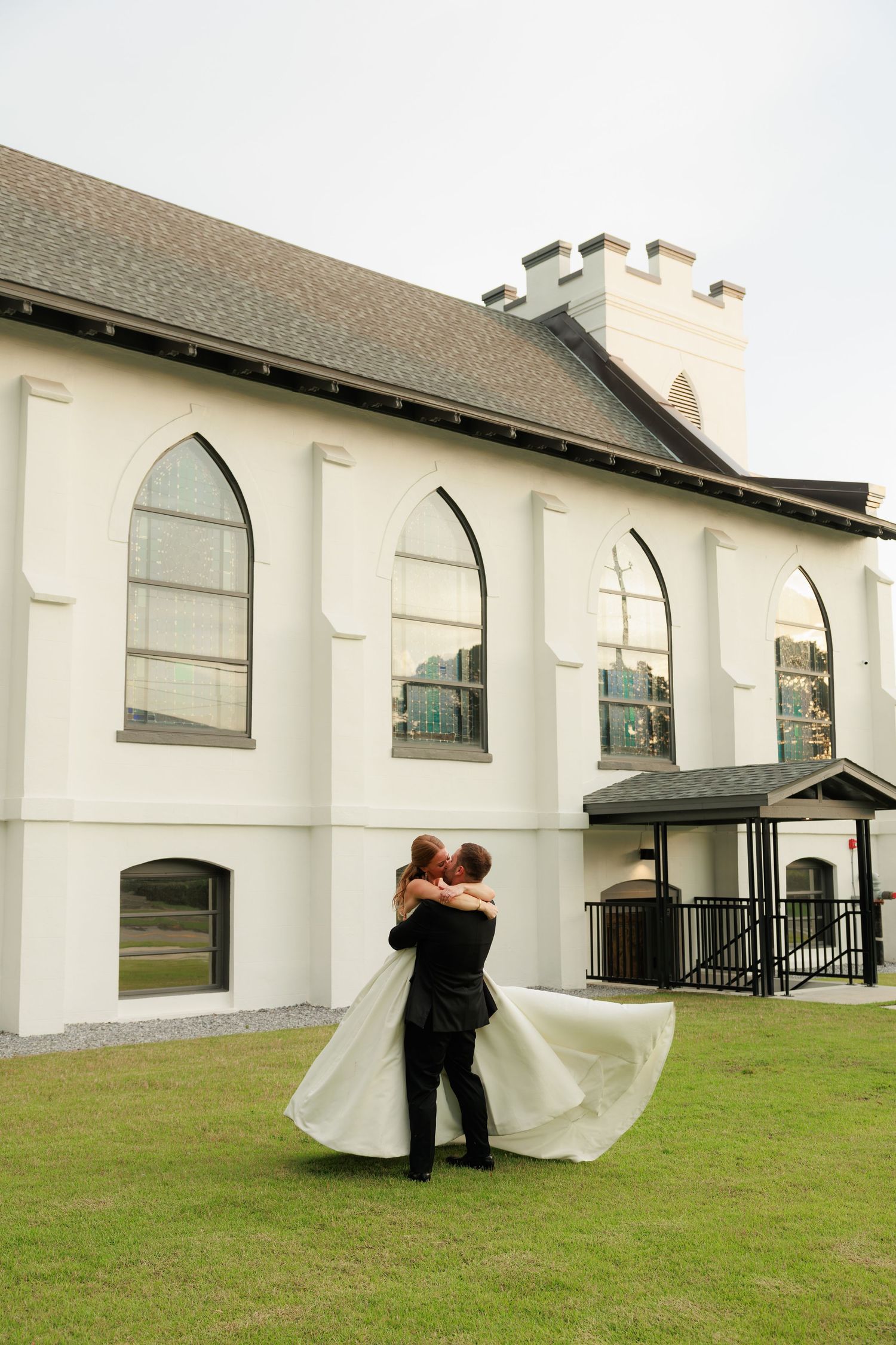 Groom lifts bride in a joyful spin on green lawn outside a white church, her gown flowing beautifully.