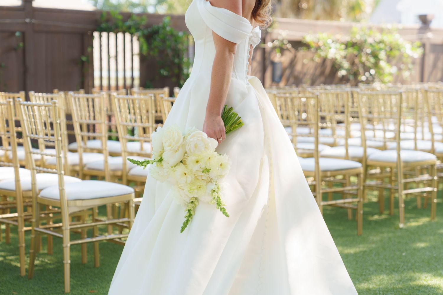 Bride in off-shoulder white gown holding white floral bouquet walks past gold chiavari chairs at outdoor wedding ceremony.
