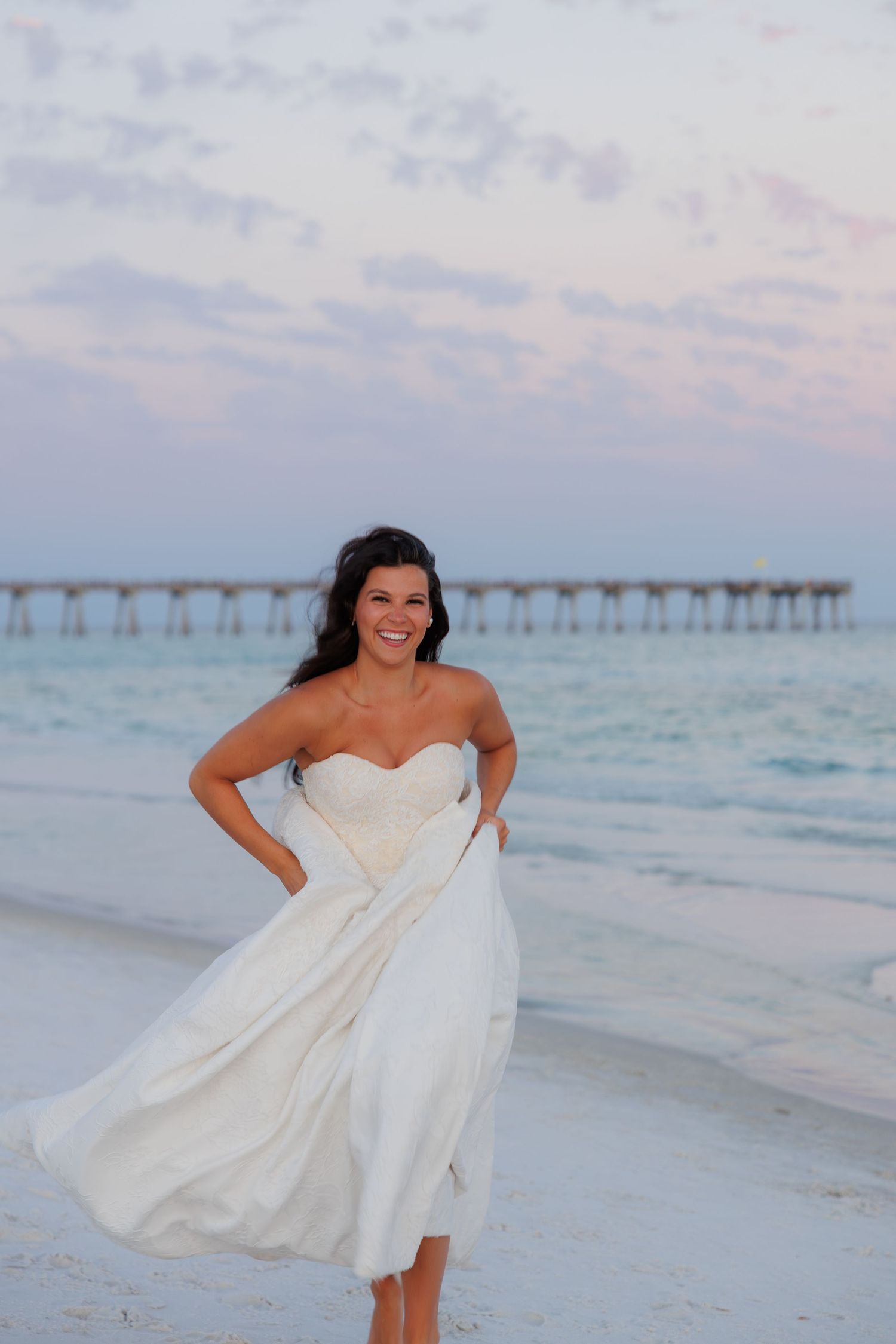 Laughing bride in strapless white gown walks on beach at dusk with pier and colorful sky behind her.