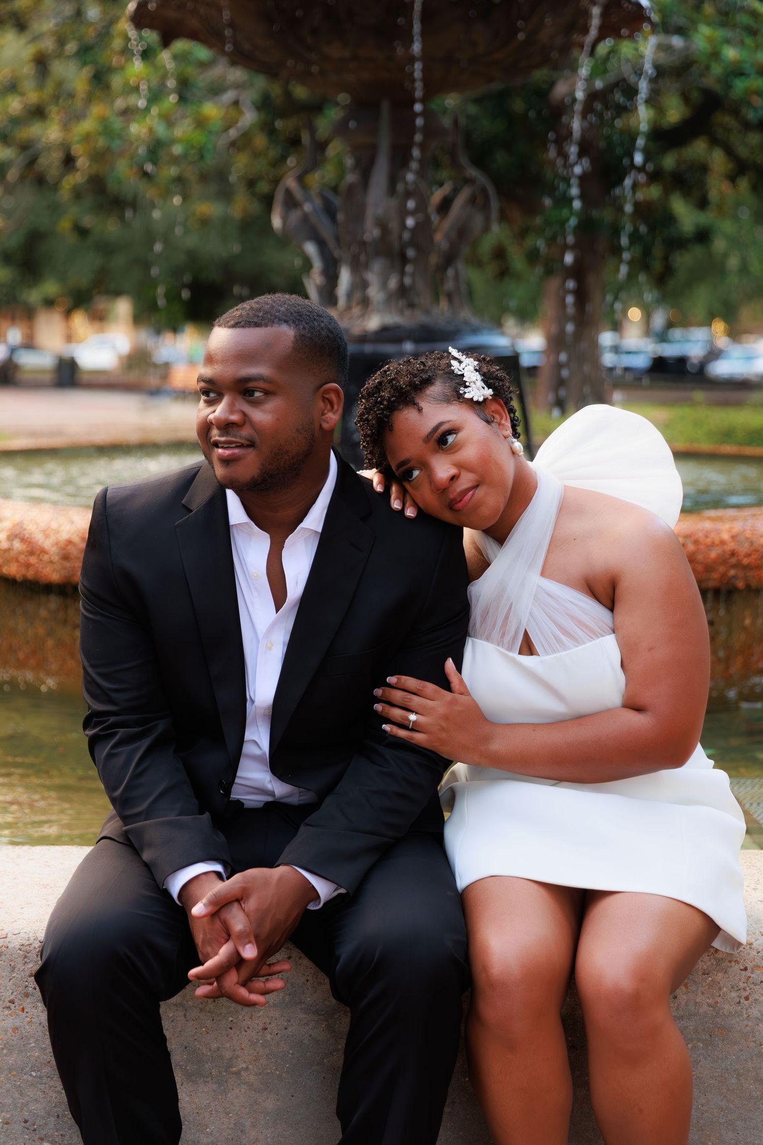 A couple in wedding attire sits by a fountain, the bride in white leaning on the groom in a black suit.