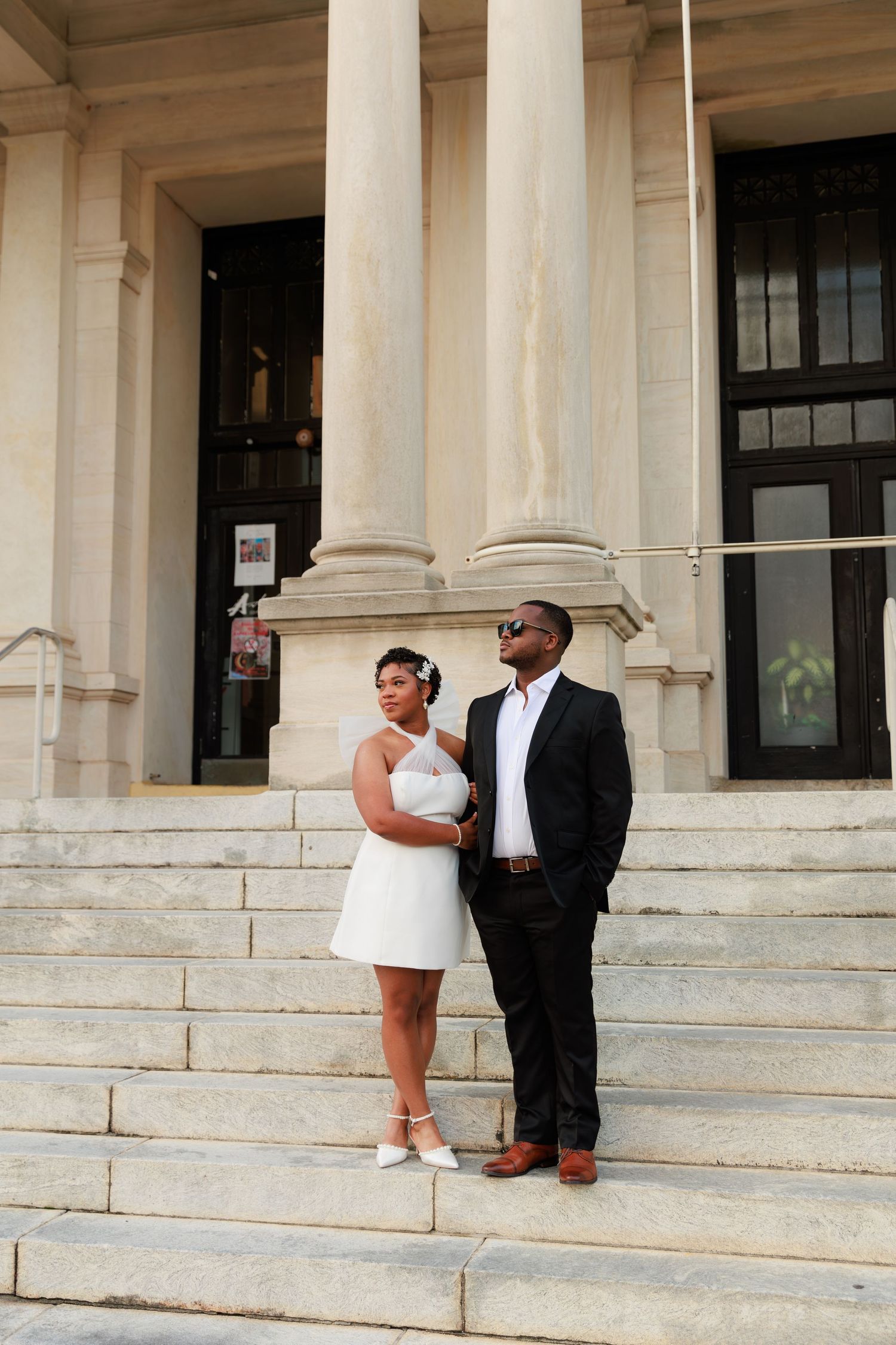 Couple in wedding attire poses on grand stone steps outside a classical building with tall columns.