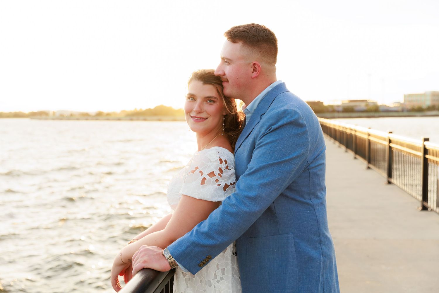 Couple in wedding attire sharing a romantic moment on a waterfront pier at golden hour sunset.