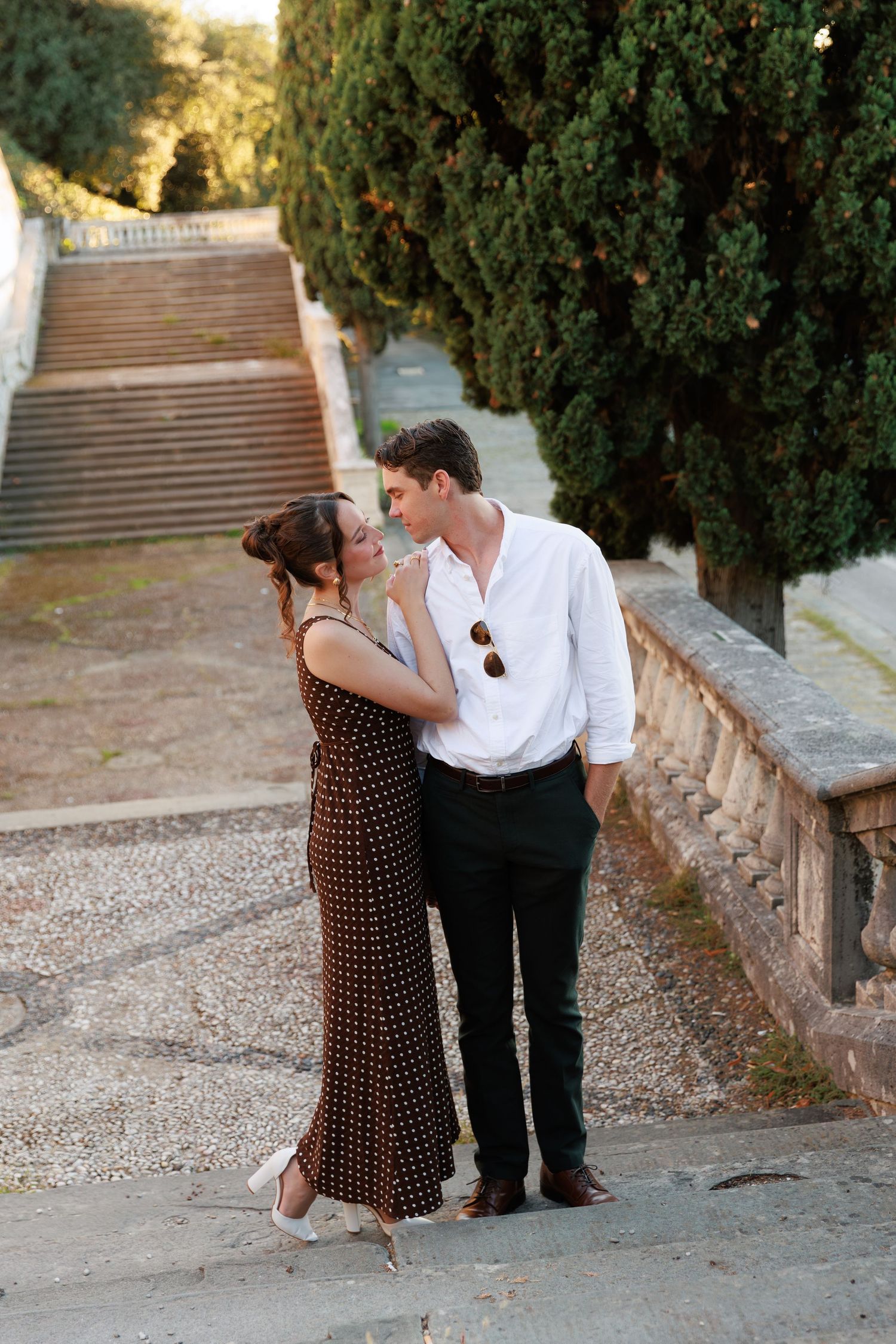 Romantic couple embracing near stone balustrade with cypress trees, woman in brown polka dot dress and white heels.