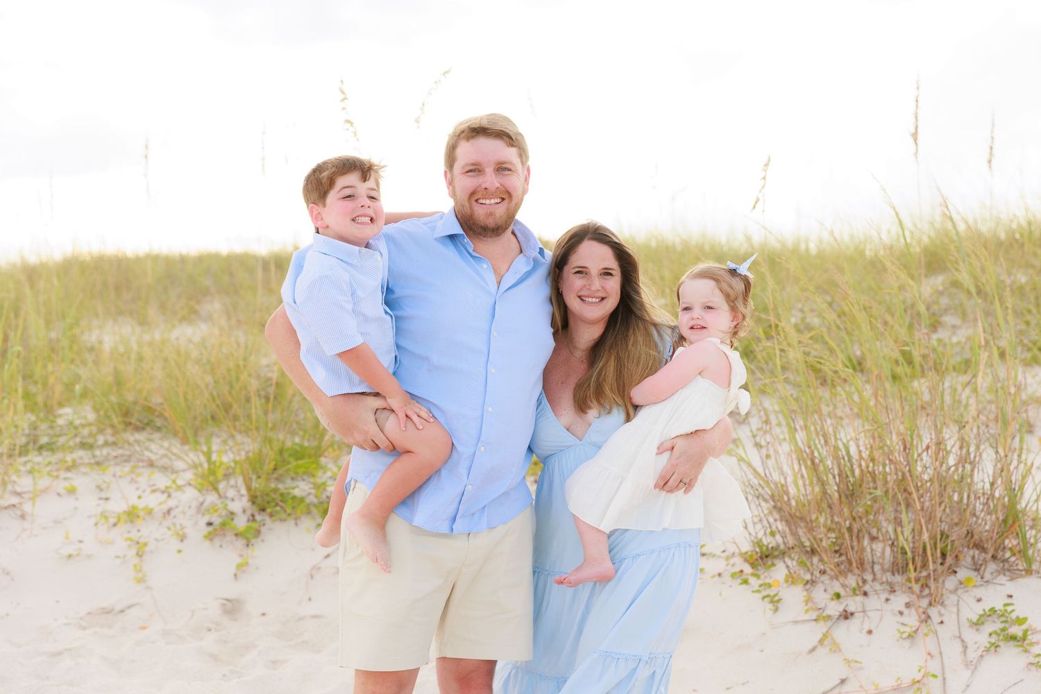 Happy family of four posing on sandy beach with sea oats, parents holding young children in coordinating blue and white outfits.