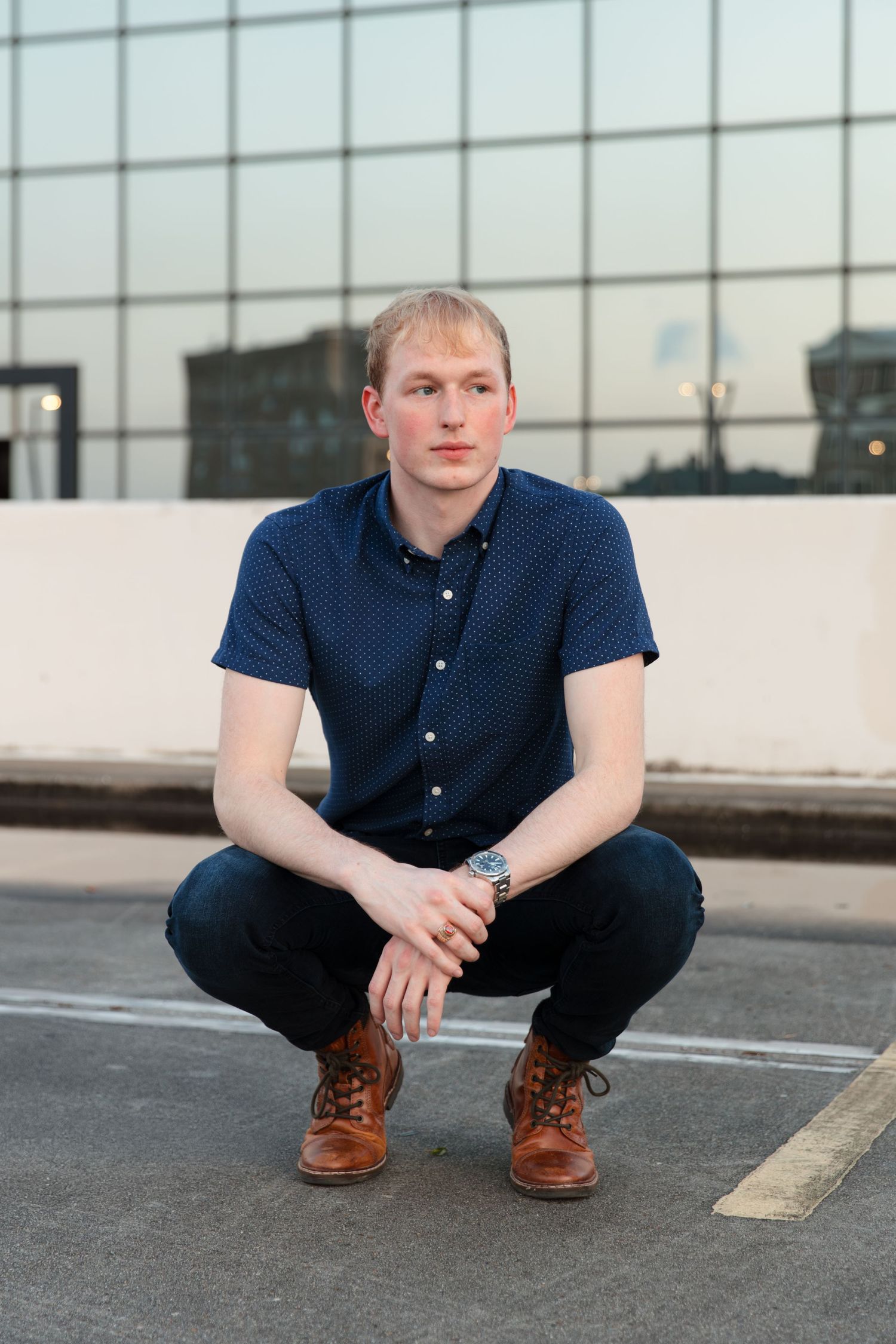 Young man in navy shirt and brown boots crouching on rooftop with glass building backdrop.