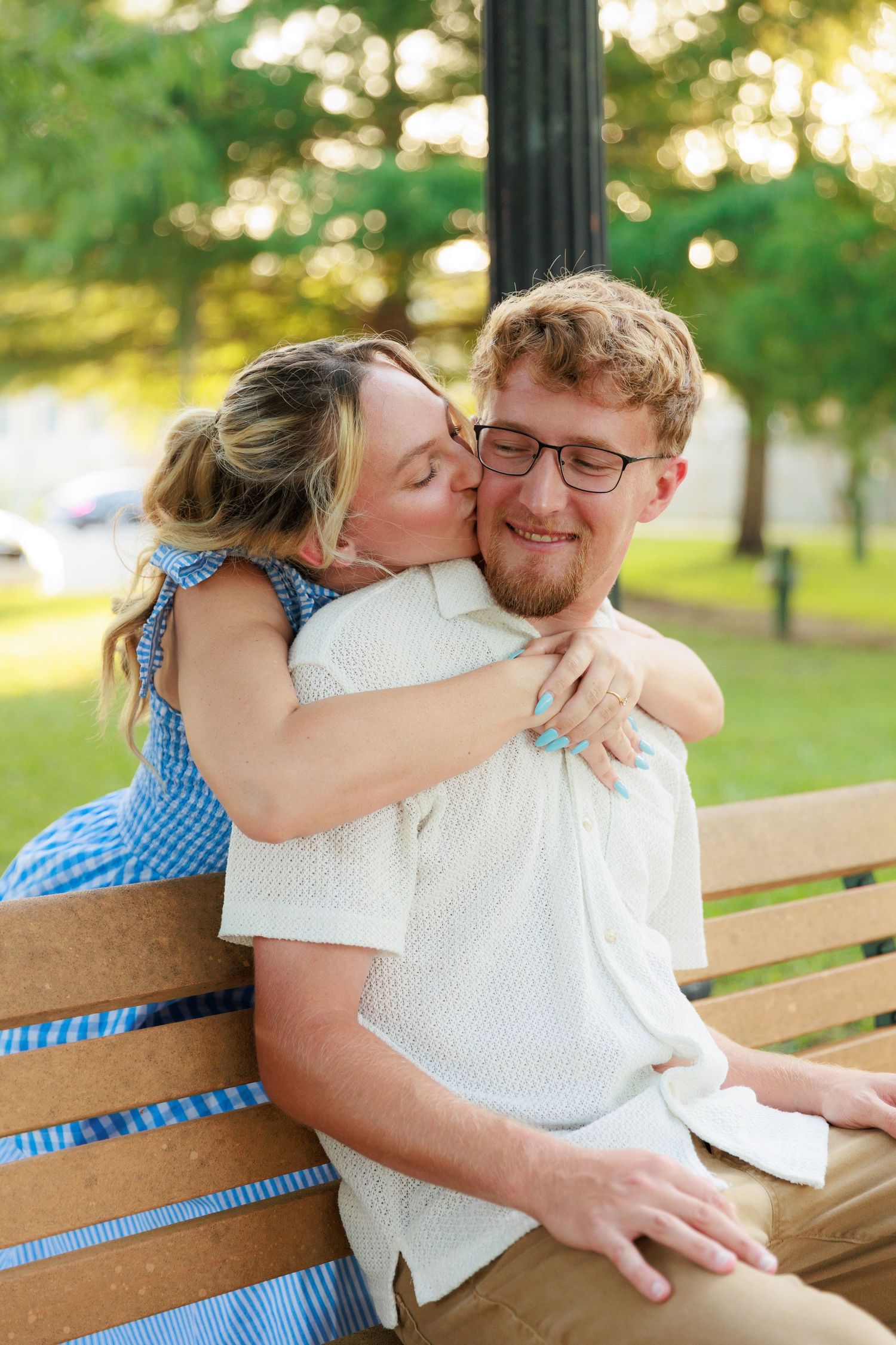 Woman in blue dress hugging and kissing man in glasses on park bench outdoors.