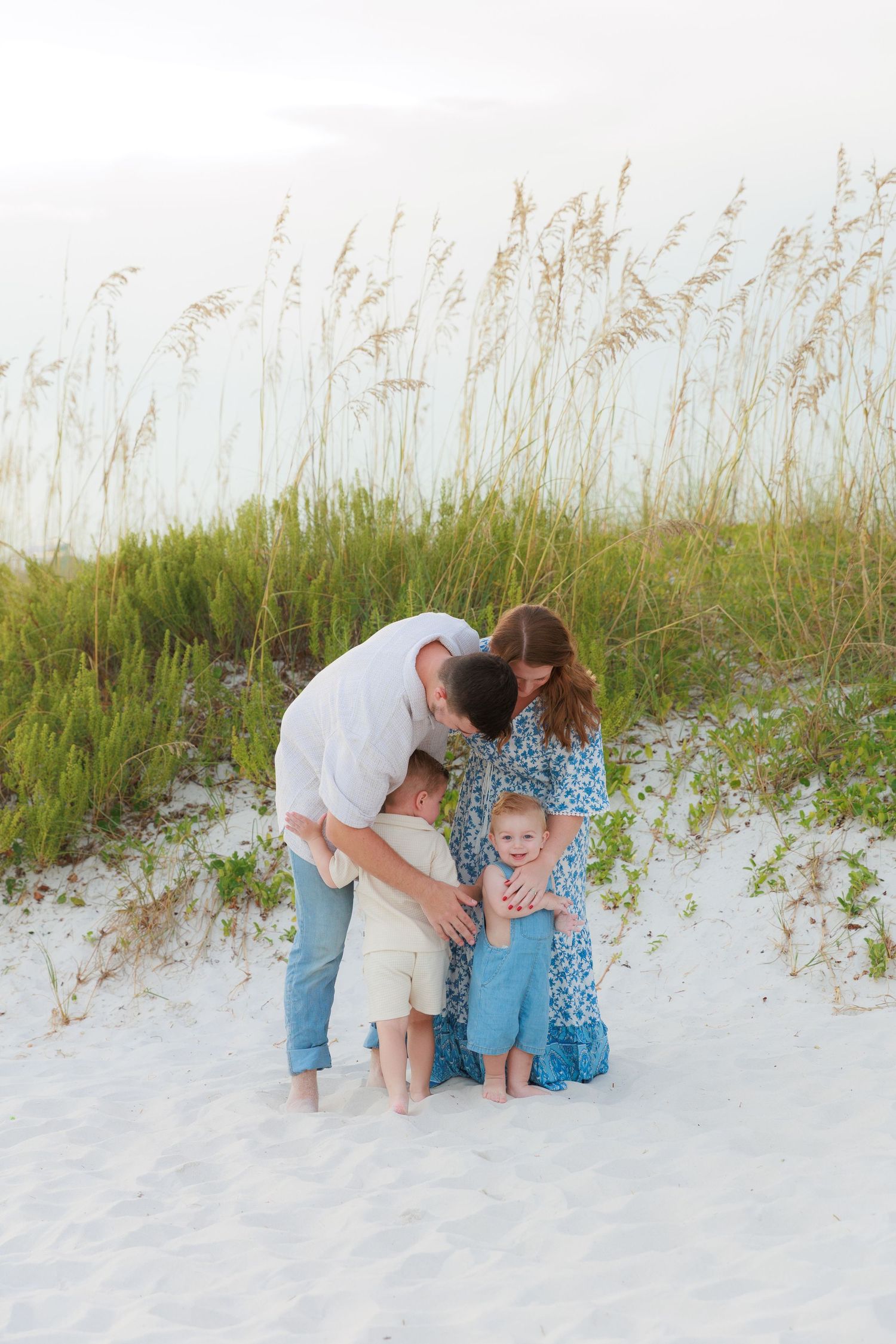 Family of four sharing a tender moment on white sand beach dunes with sea oats in background.