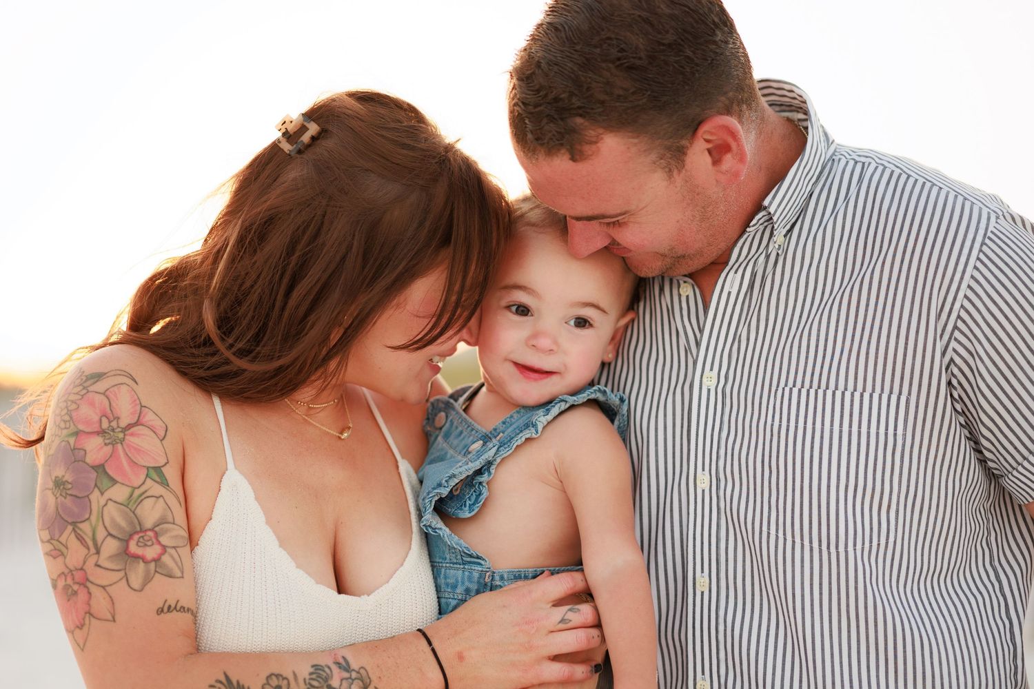 Happy family portrait of parents with tattooed mom and dad lovingly holding smiling toddler at sunset beach.