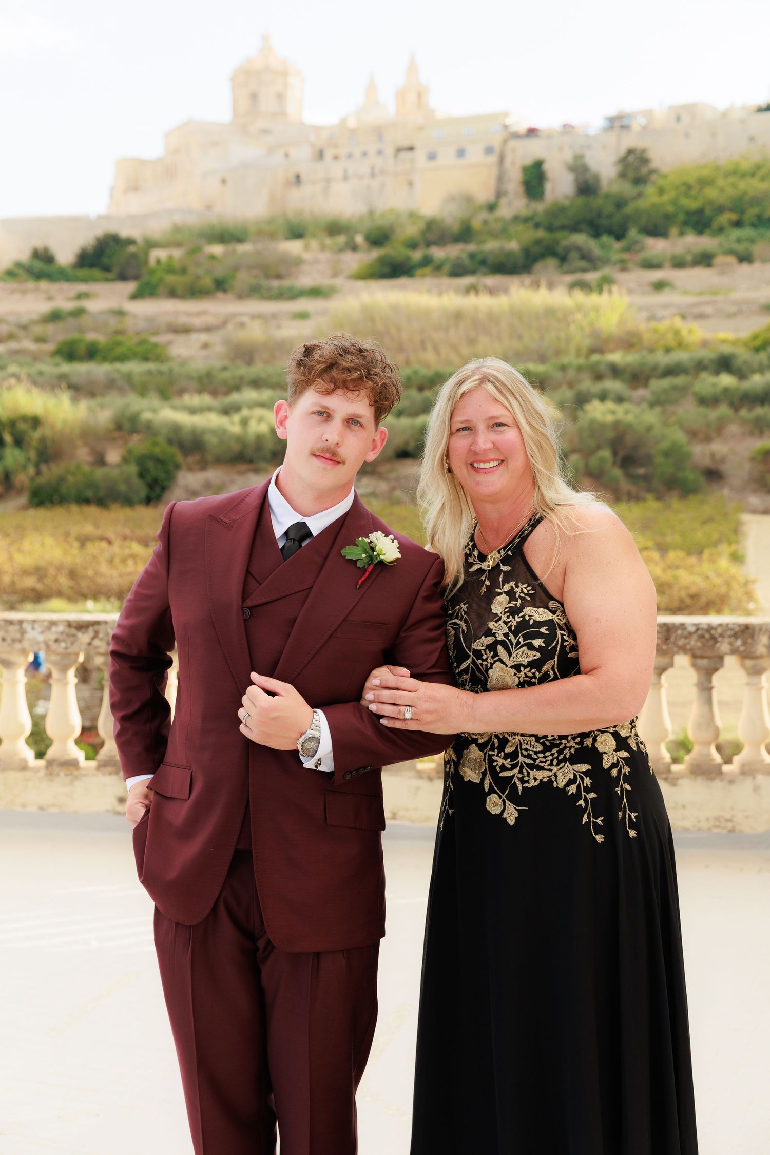 A formal portrait of two people in formal attire, one in a burgundy suit and one in a black and gold dress, outdoors.