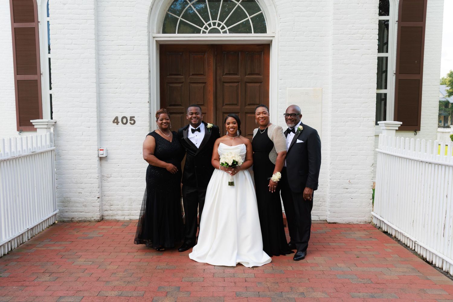 Wedding party gathers for informal photos outside a white church building with colonial architecture.