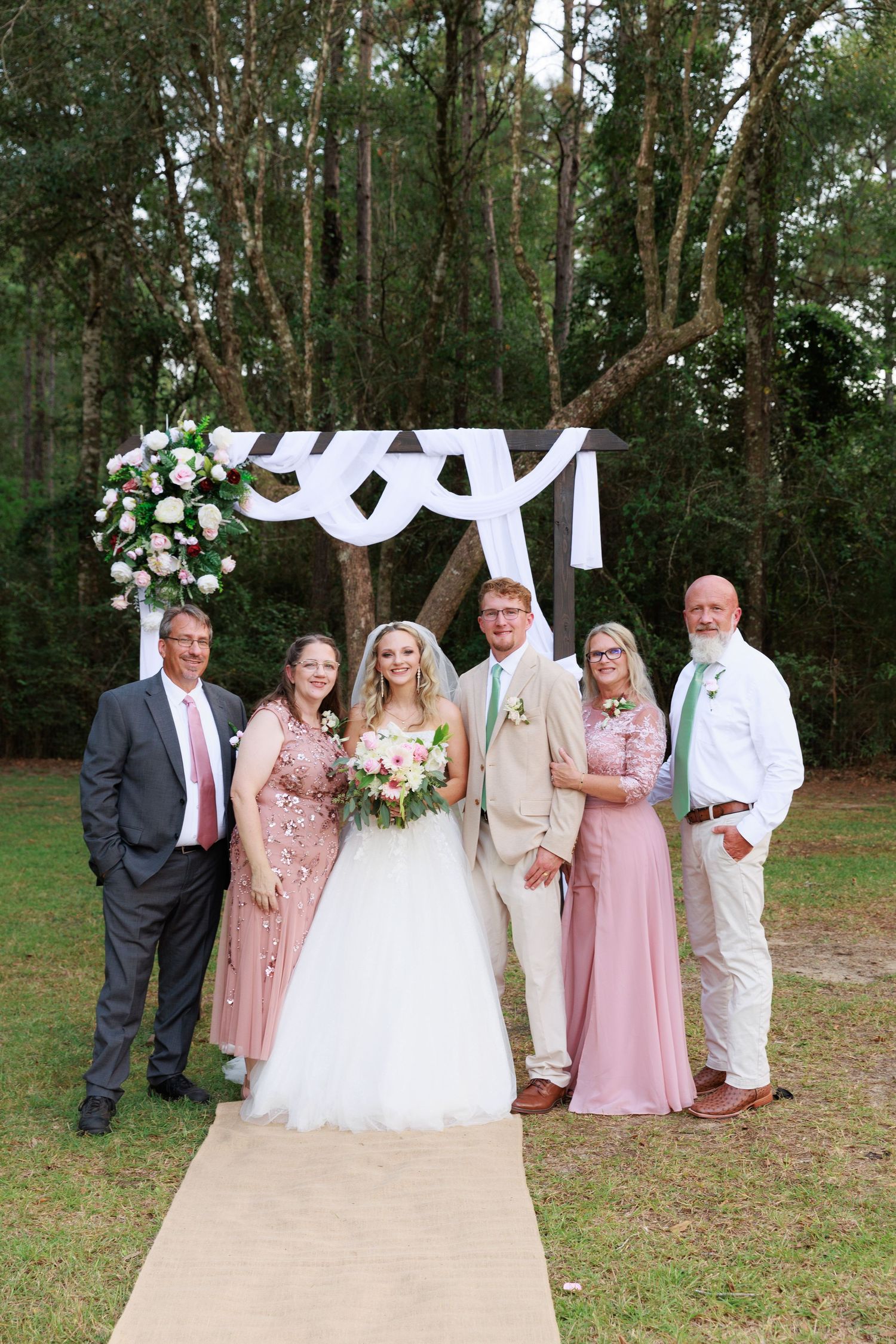 A wedding party in pink and white attire poses together outdoors in a wooded setting.