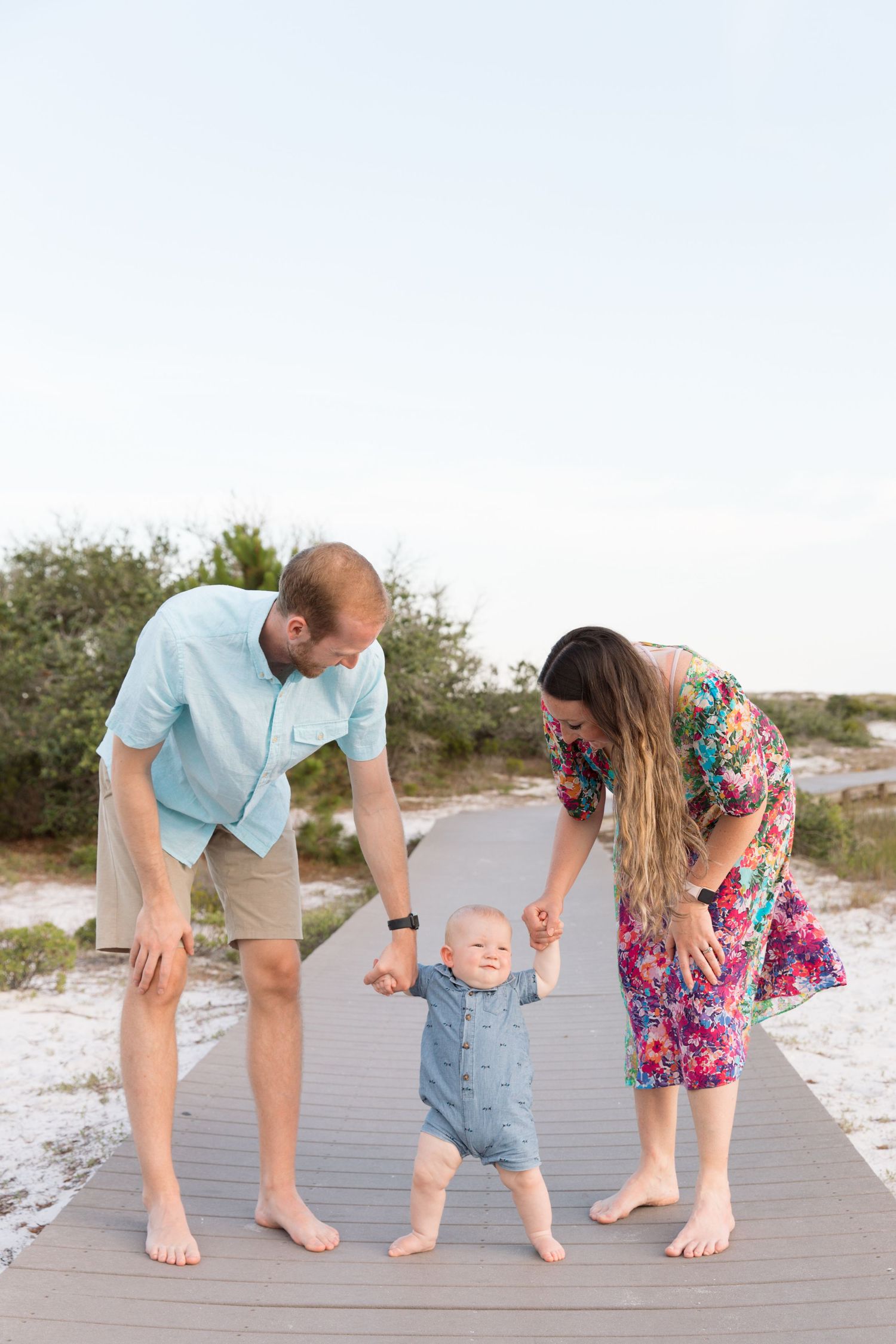 A young family helps their baby take first steps on a beachside boardwalk, parents smiling lovingly at their child.