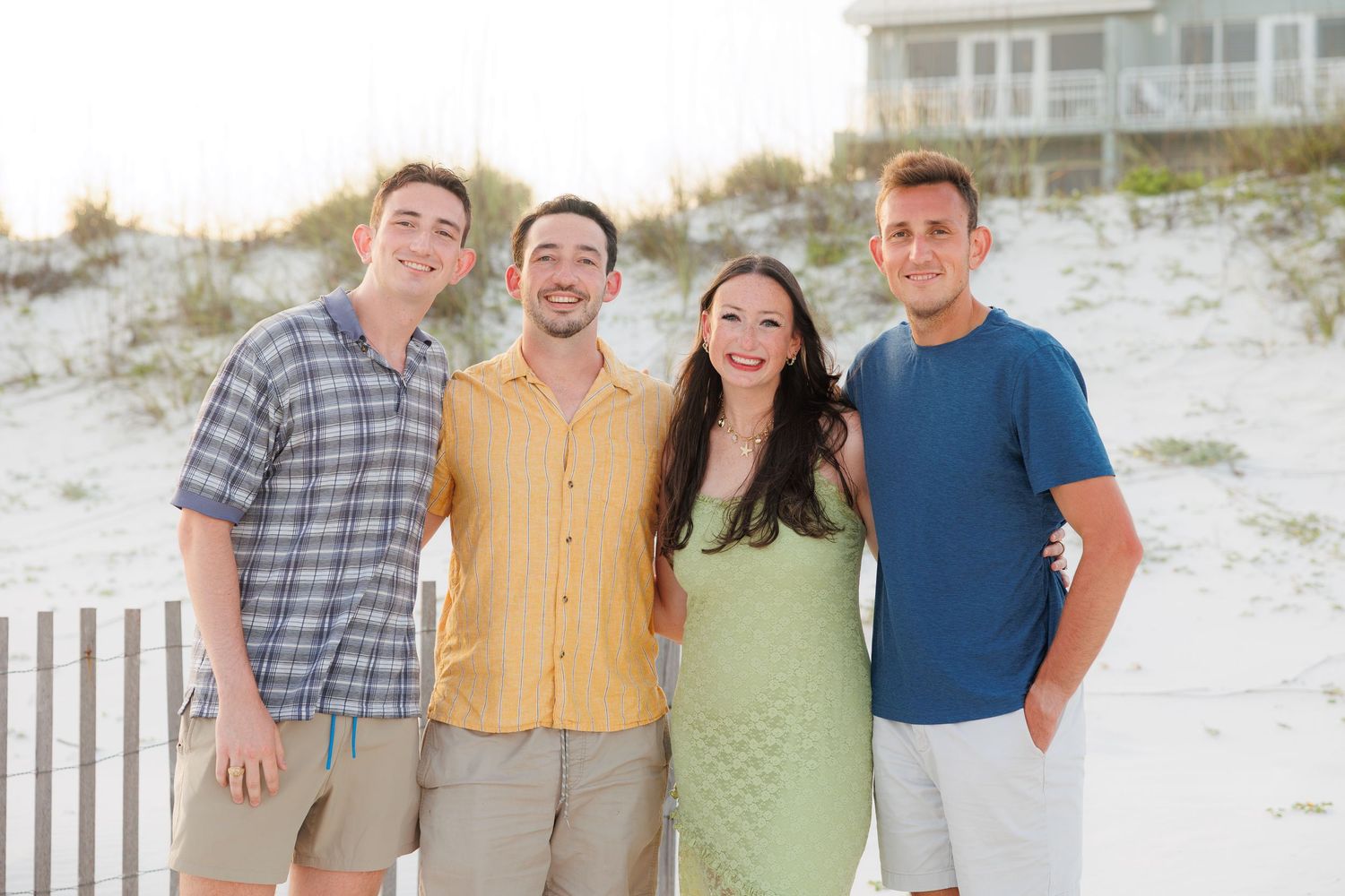 Four smiling friends pose together on a sunny beach with white sand dunes and a house in the background.