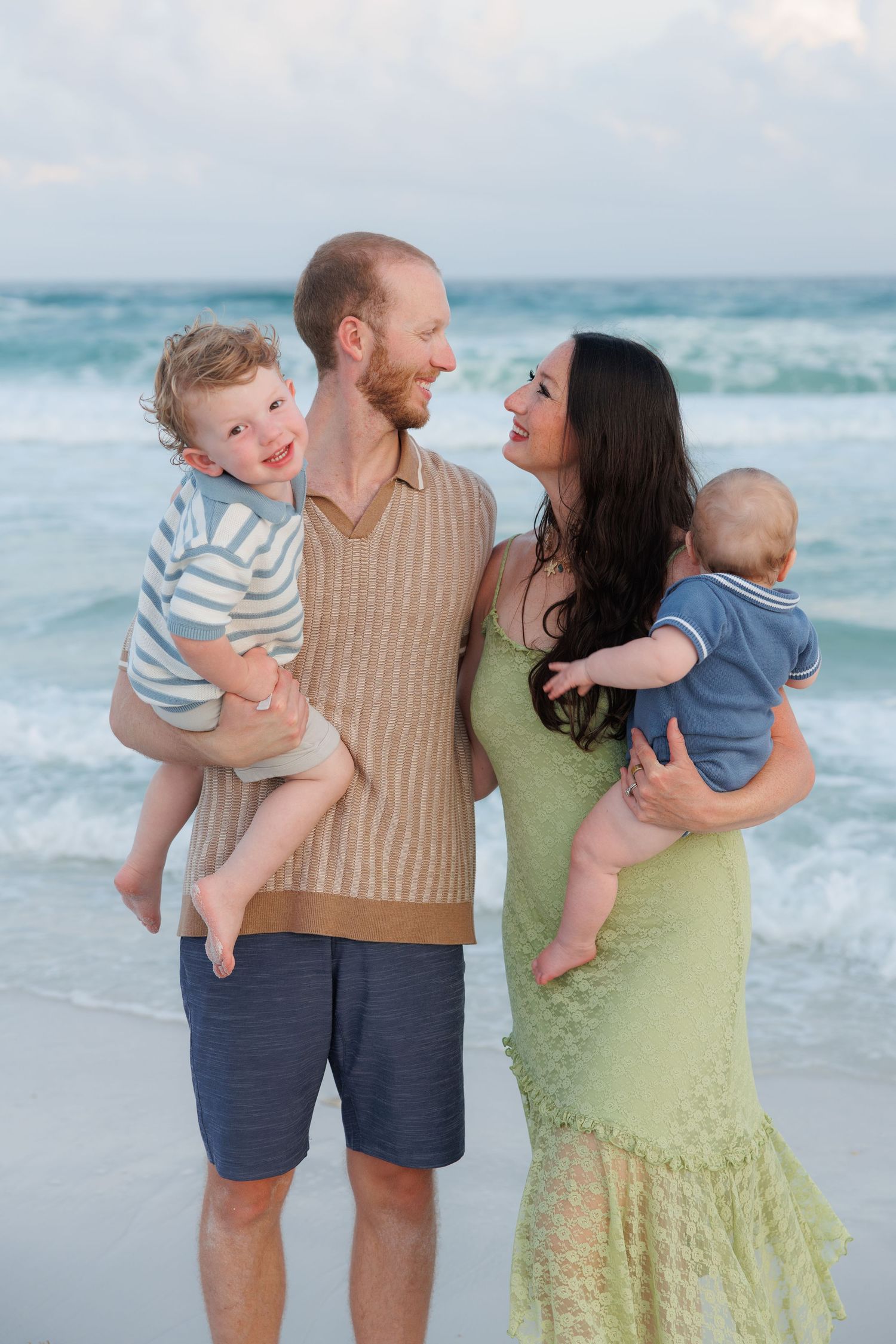 Happy family of four with two young boys posing together on a beach with turquoise ocean waves behind them.