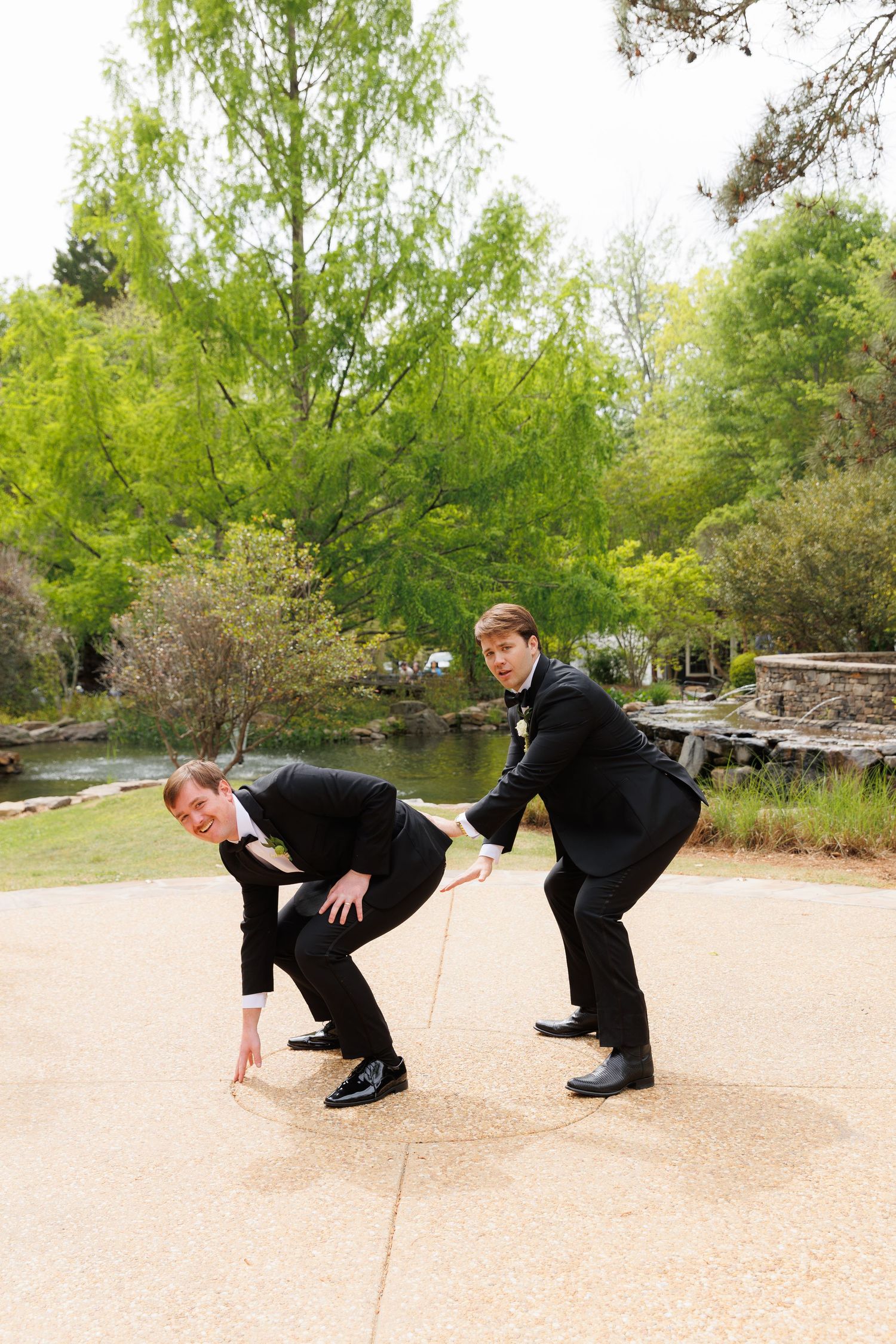 Two groomsmen in black suits pose playfully outdoors near a pond with lush green trees in background.