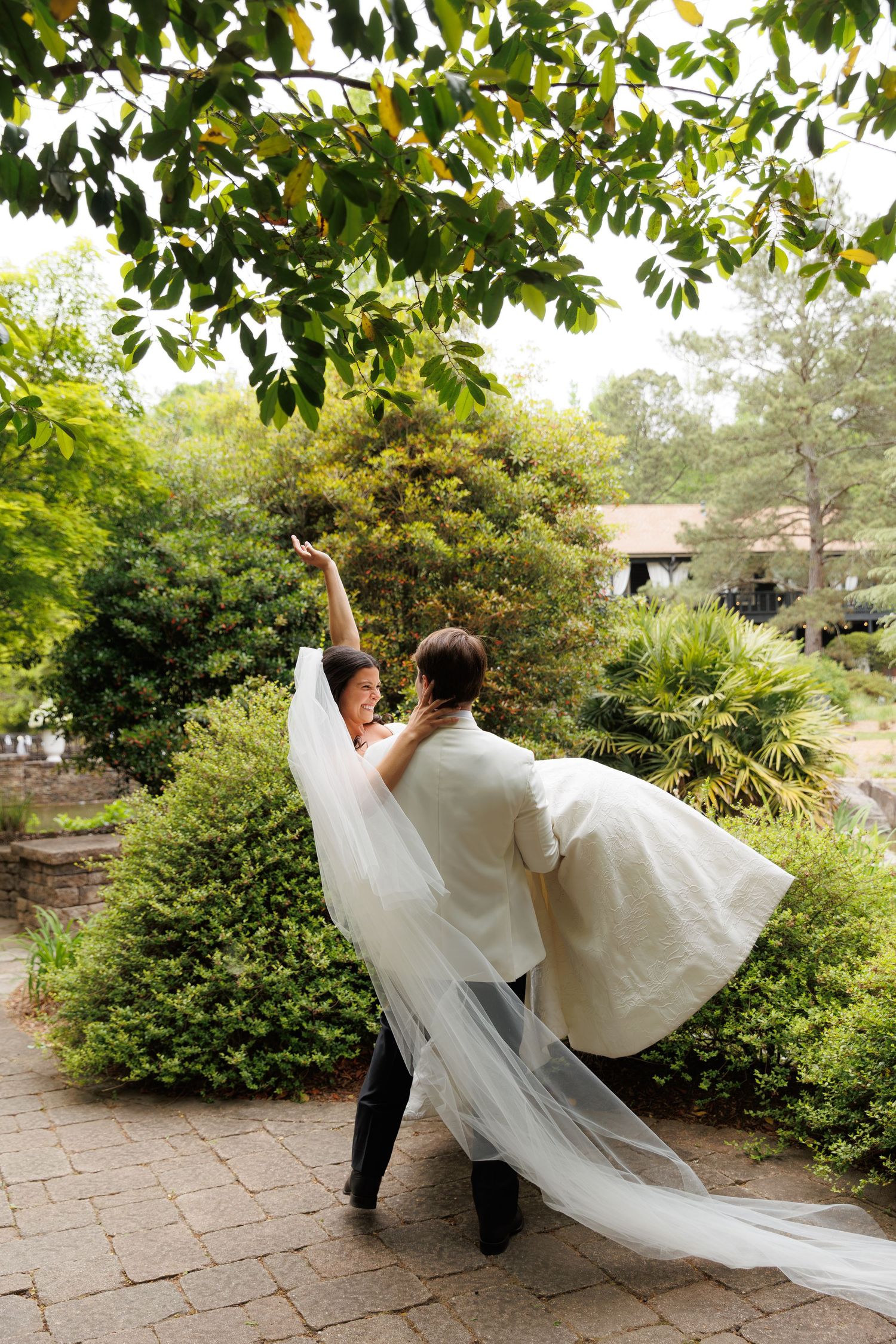 Groom lifts bride in flowing white gown and veil amid lush green garden surroundings on their wedding day.