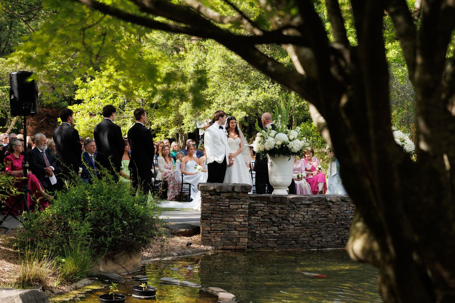 Outdoor garden wedding ceremony with couple at altar, lush green trees, stone bridge, and seated guests in elegant setting.