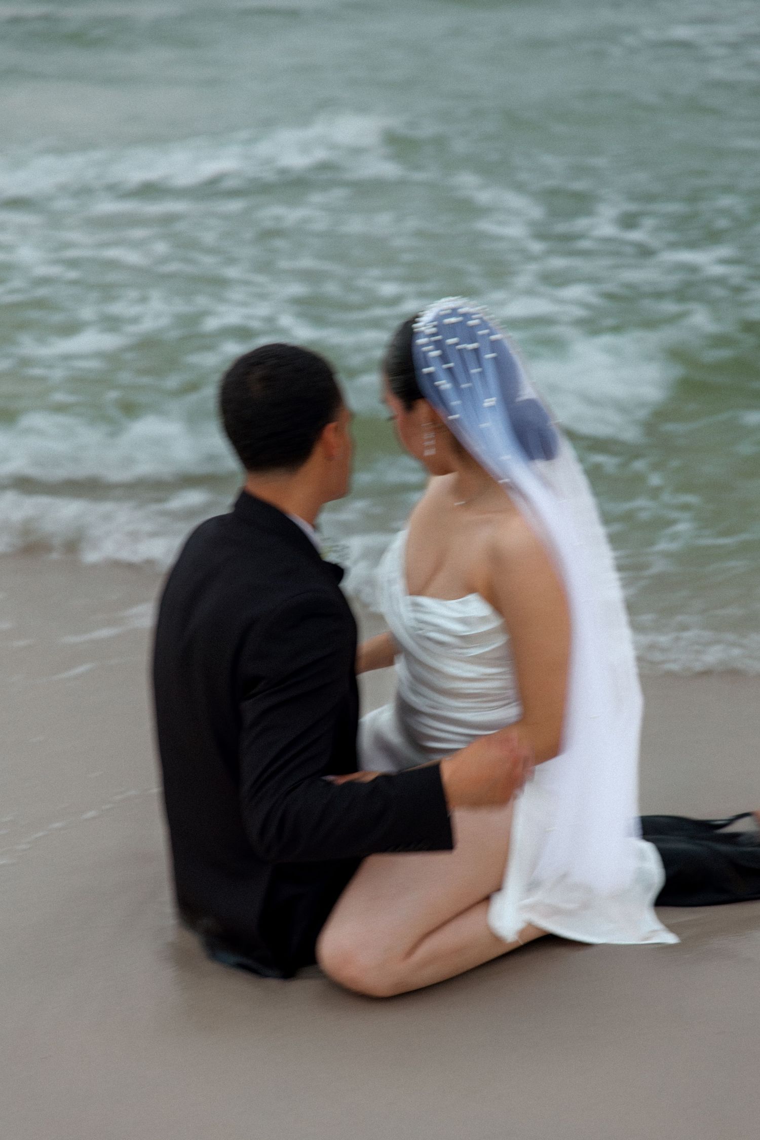 Couple sits together at water's edge during beach wedding photoshoot.