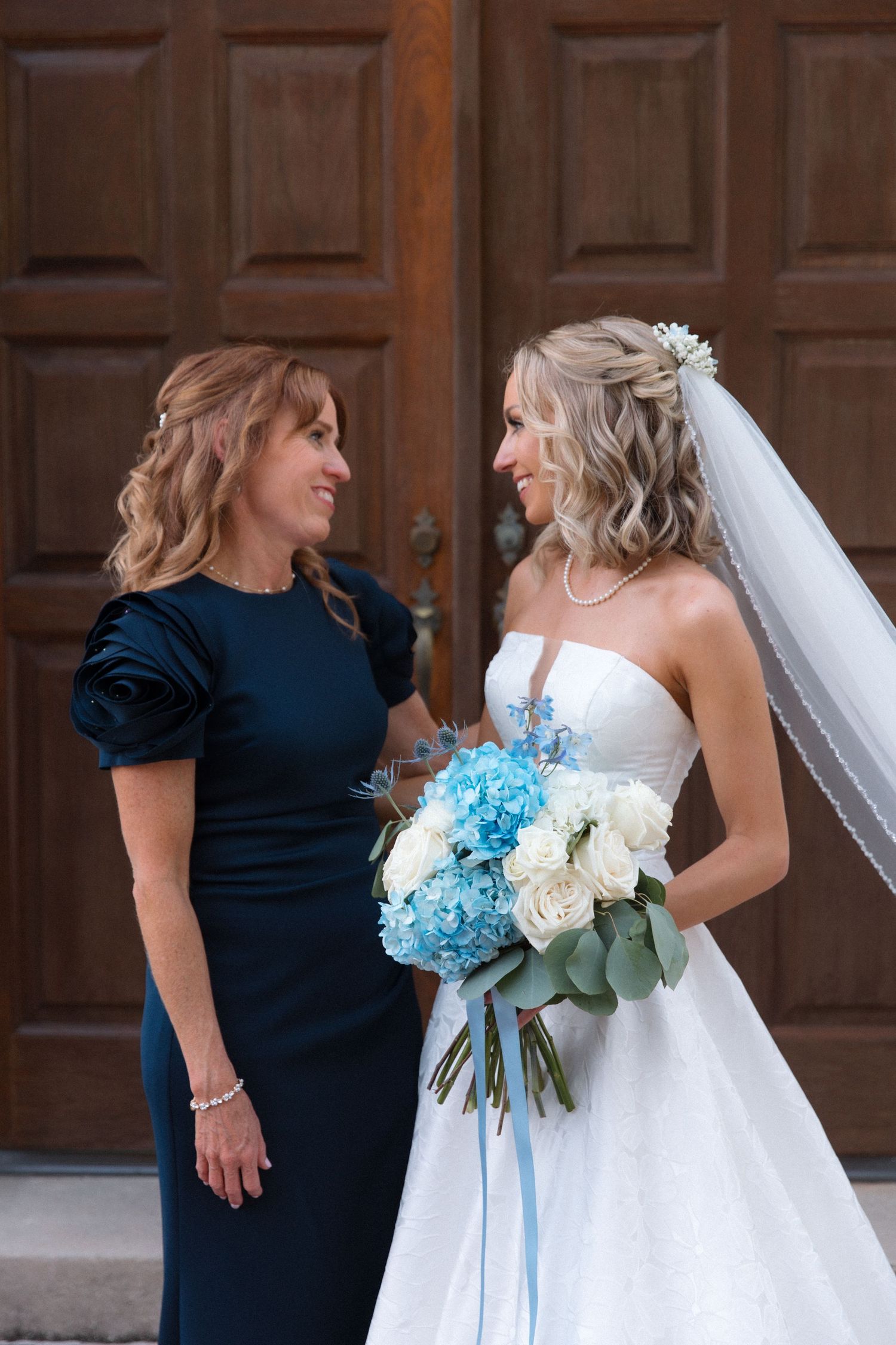 Beautiful bridal moment captured with navy-dressed companion in front of wooden door.