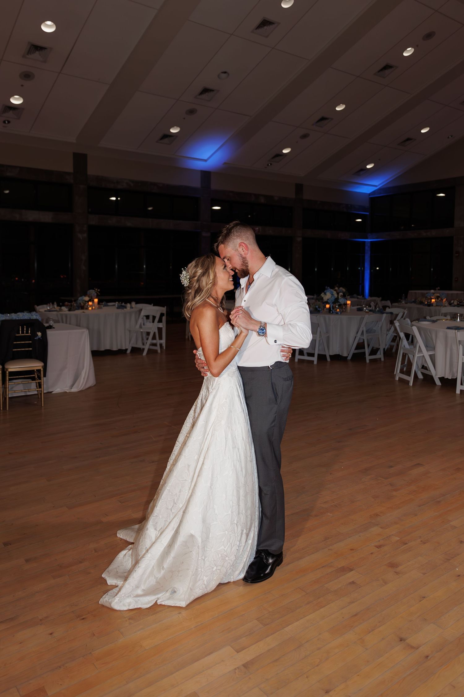 Bride and groom slow dancing in elegant reception hall with blue lighting and white table settings.
