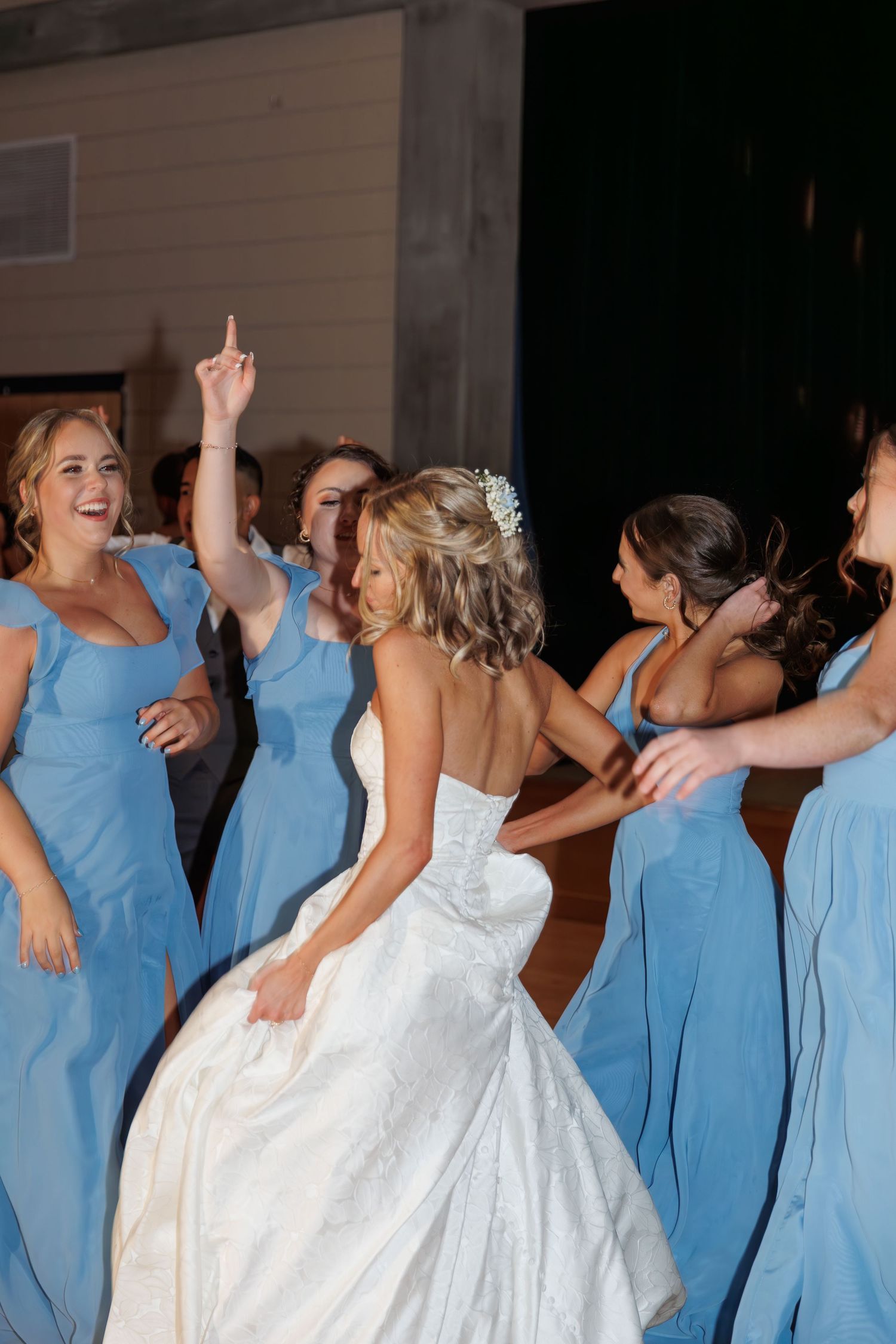 A bride in white gown dances joyfully with bridesmaids wearing light blue dresses.