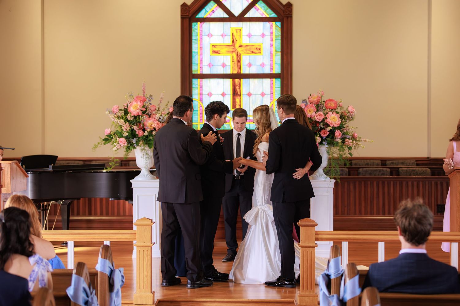 Wedding ceremony in progress at the altar beneath a stained glass window with a cross and pink flower arrangements.