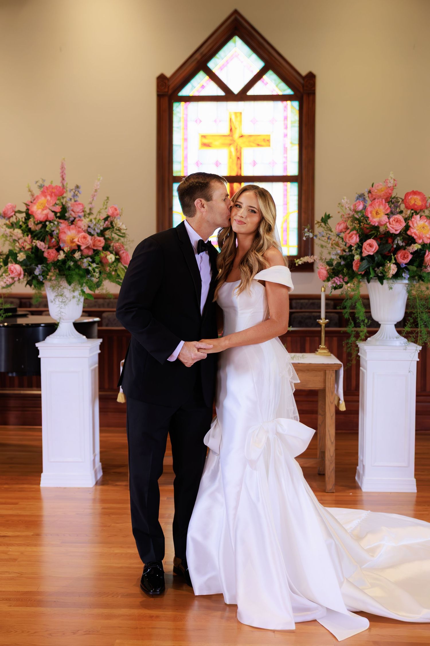 A bride and groom share an intimate moment in a chapel with pink floral arrangements and a stained glass cross window.