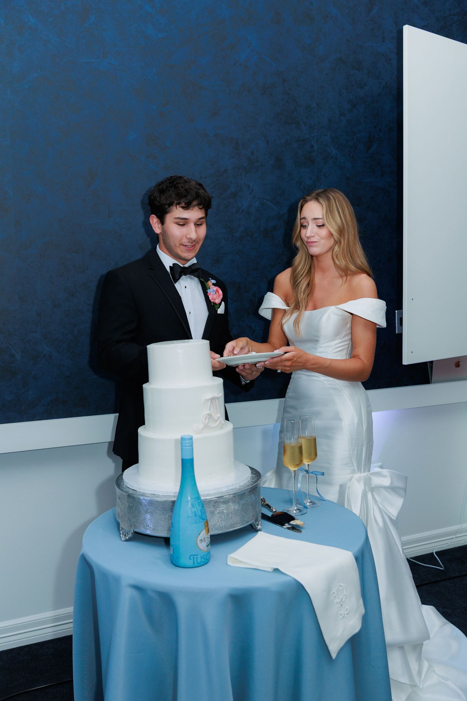 Couple in formal attire cutting their elegant white wedding cake together at reception with blue decor accents.