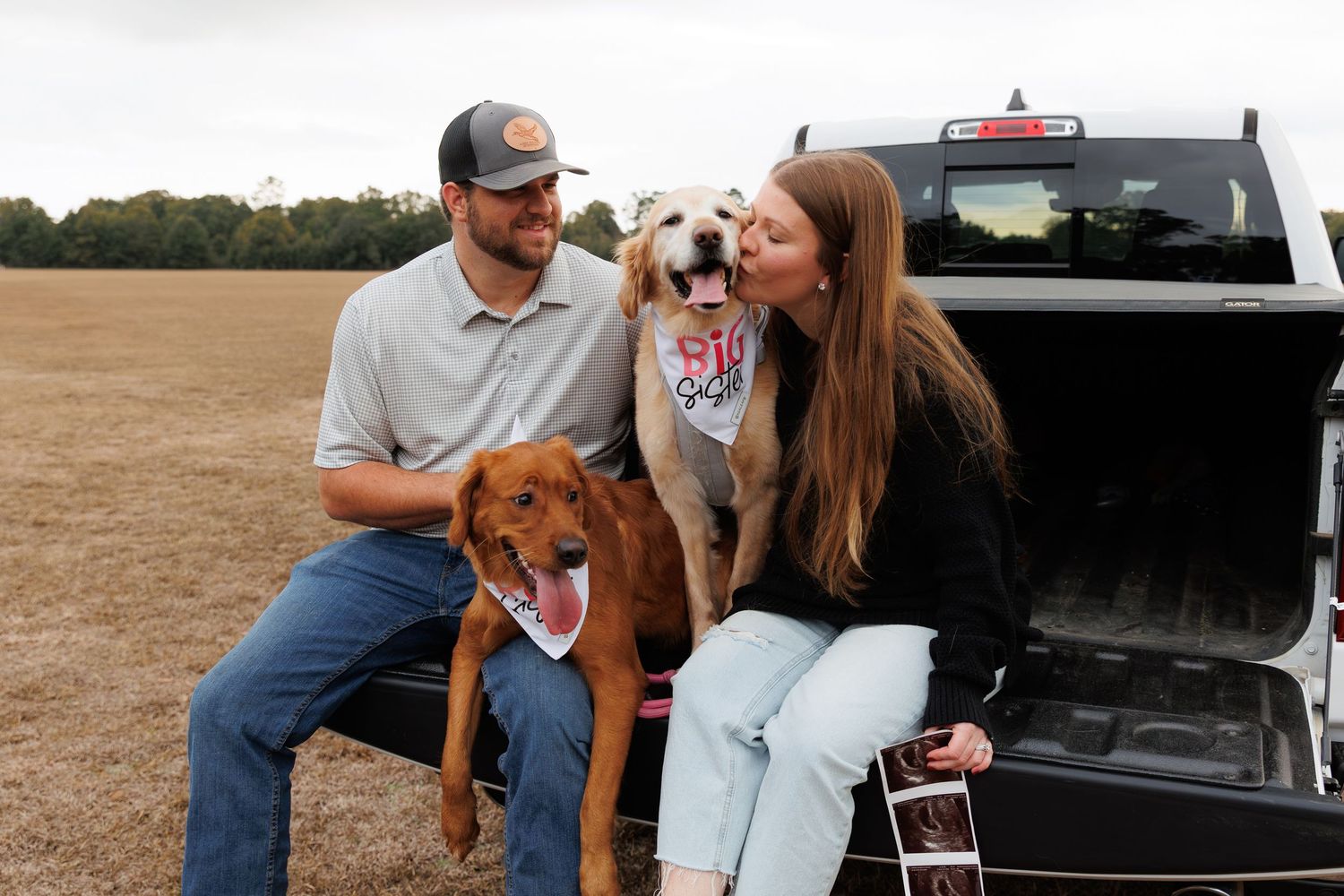 A couple sits in the back of a pickup truck with their two dogs wearing bandanas in an open field.