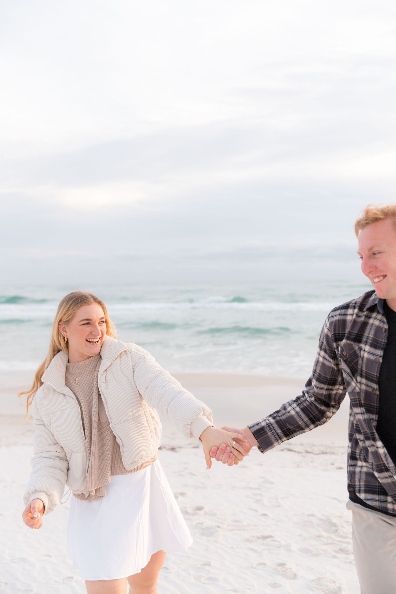 Couple in casual attire holding hands and enjoying a romantic moment on beach with ocean backdrop.