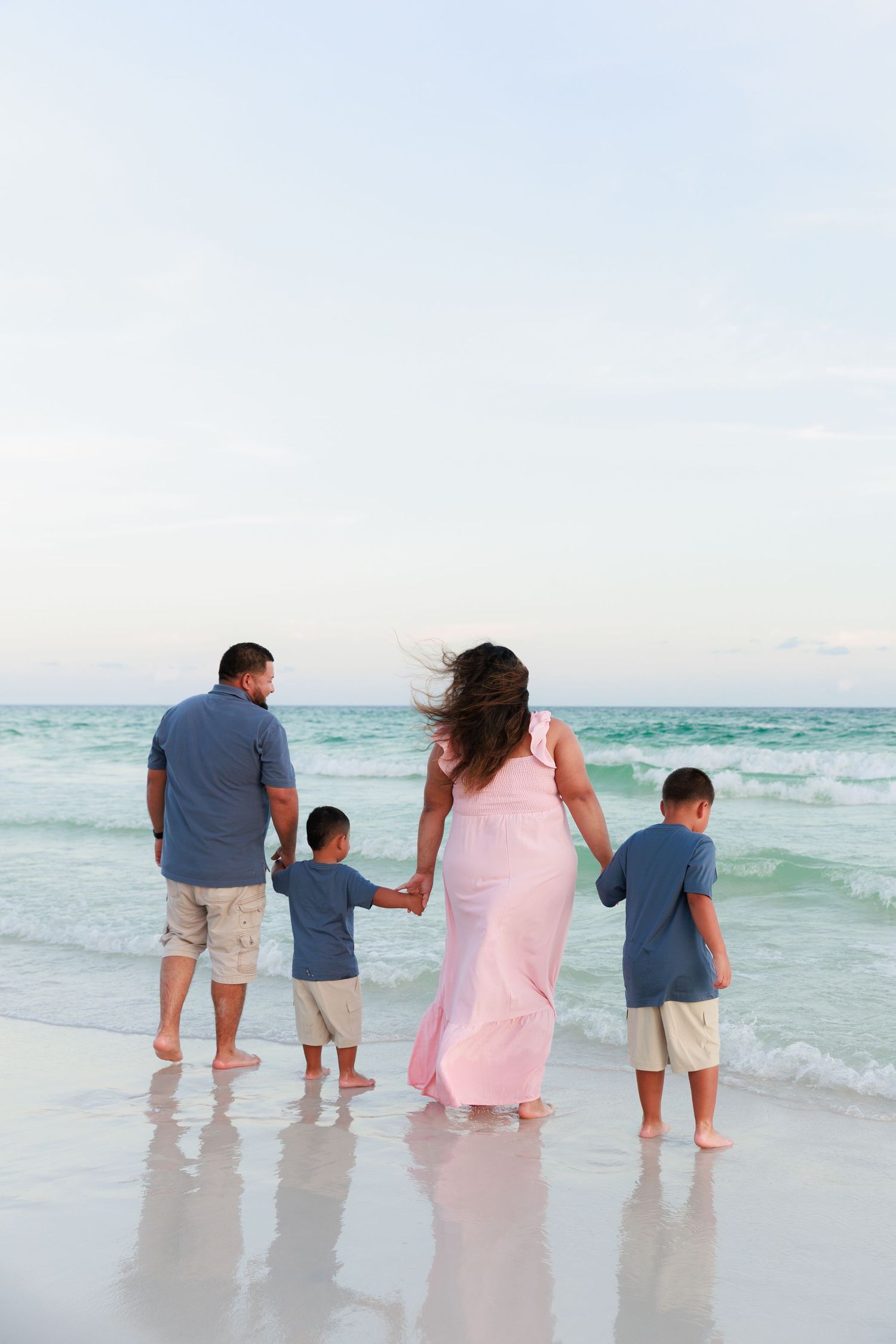 Family stands together at the edge of turquoise ocean waters on a white sandy beach during sunset.
