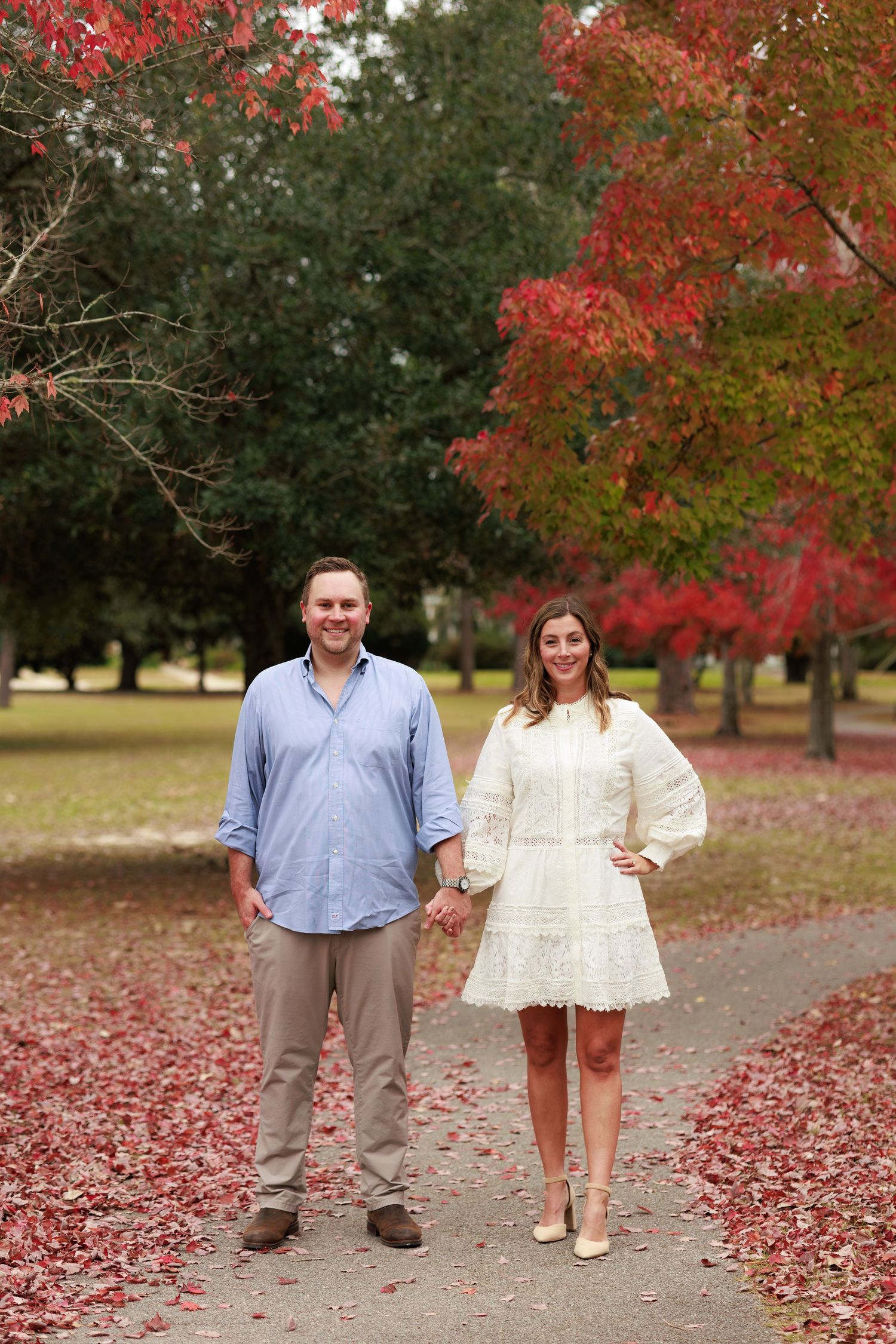A couple stands together on a carpet of fallen red autumn leaves beneath vibrant fall foliage in a park setting.