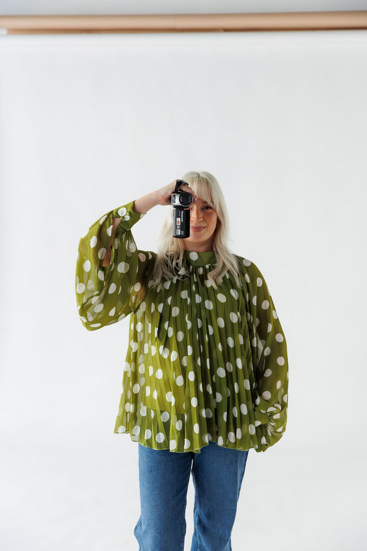 Someone wearing a green polka dot blouse and jeans holds up a camera to take a photo against a white background.