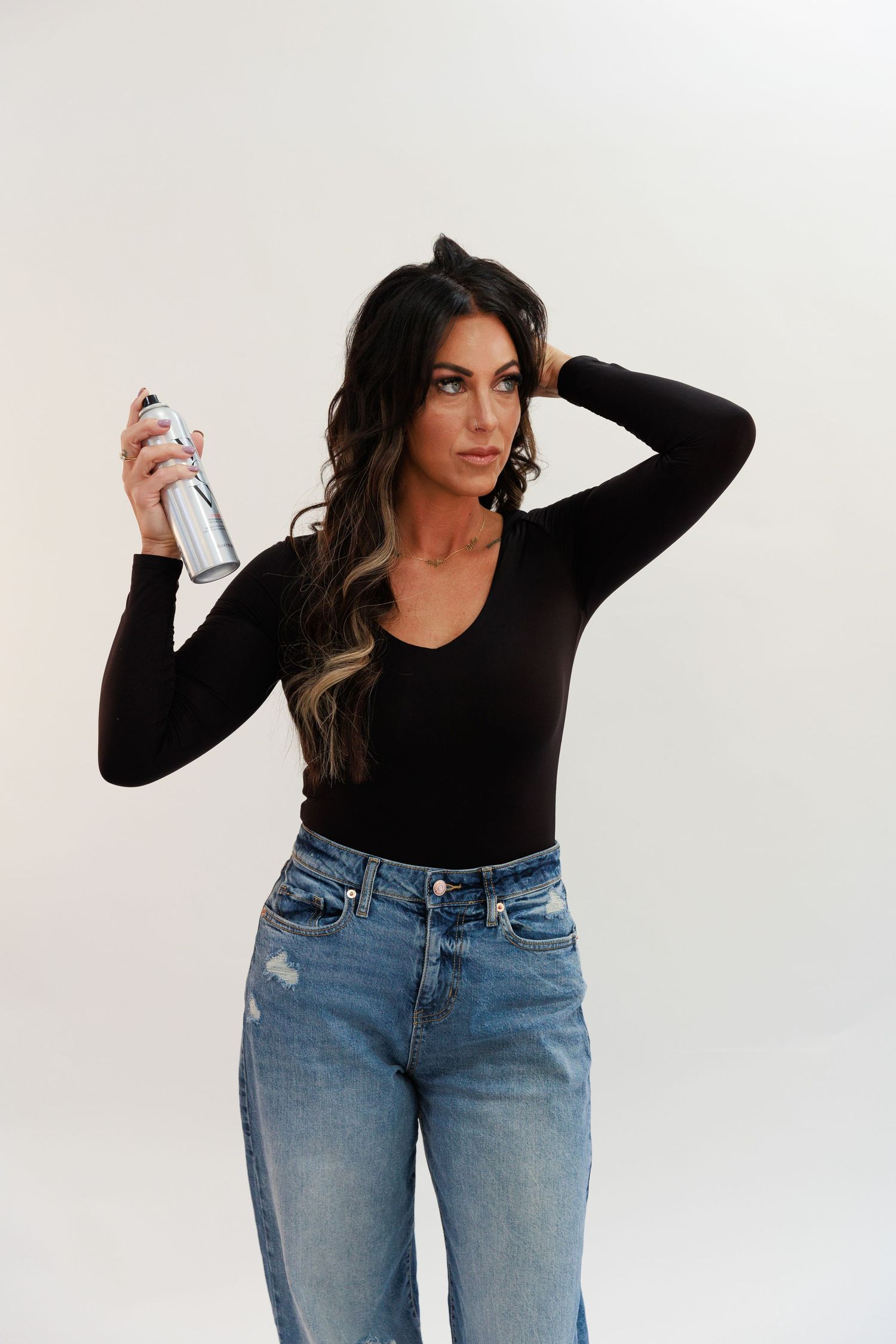 Model in black fitted top and light wash jeans holds styling product bottle.