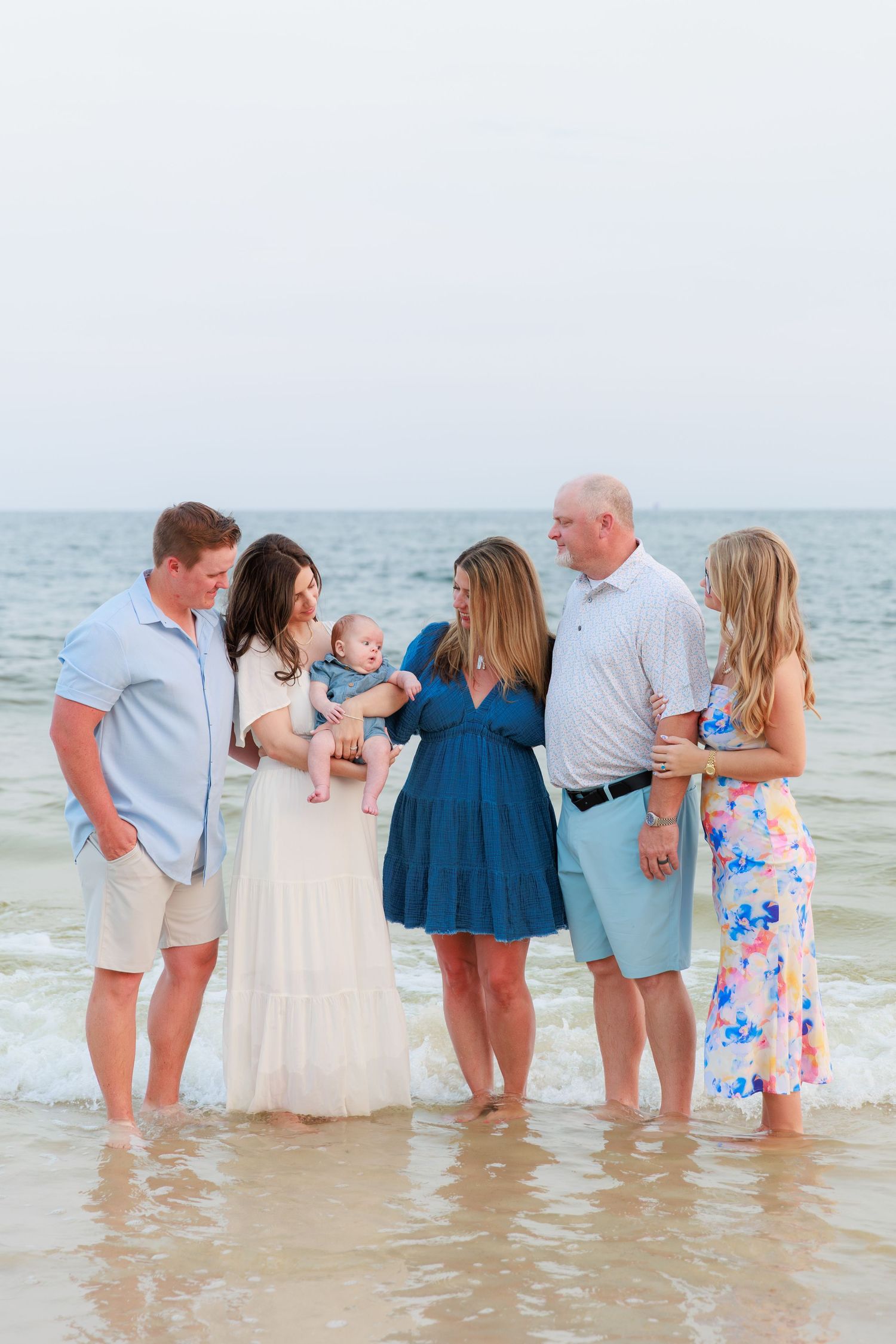Extended family of six standing in ocean waves admiring a baby held by young mother in white dress.