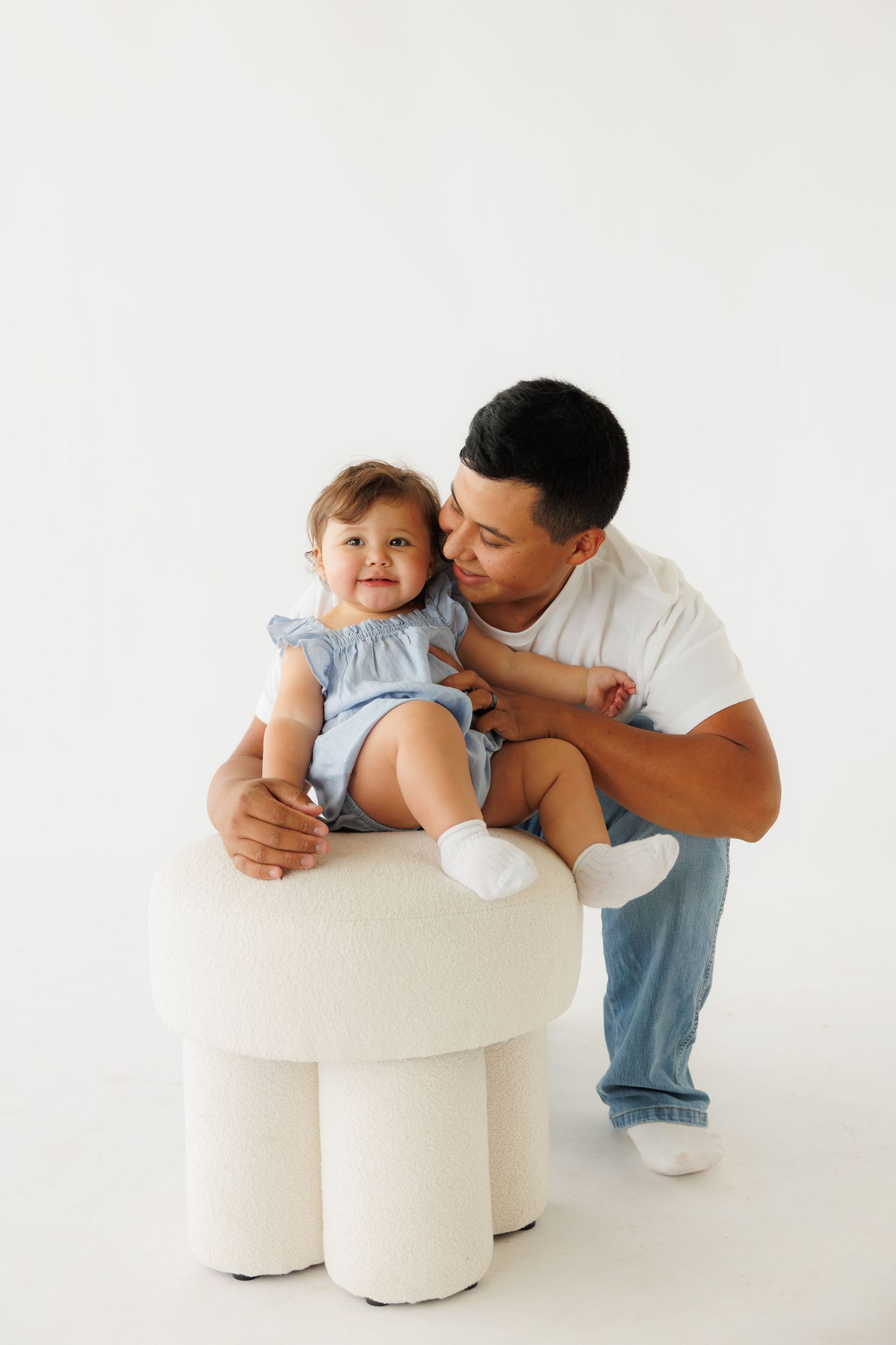 A father tenderly holds his smiling baby girl in a blue outfit on a white stool against a white background.
