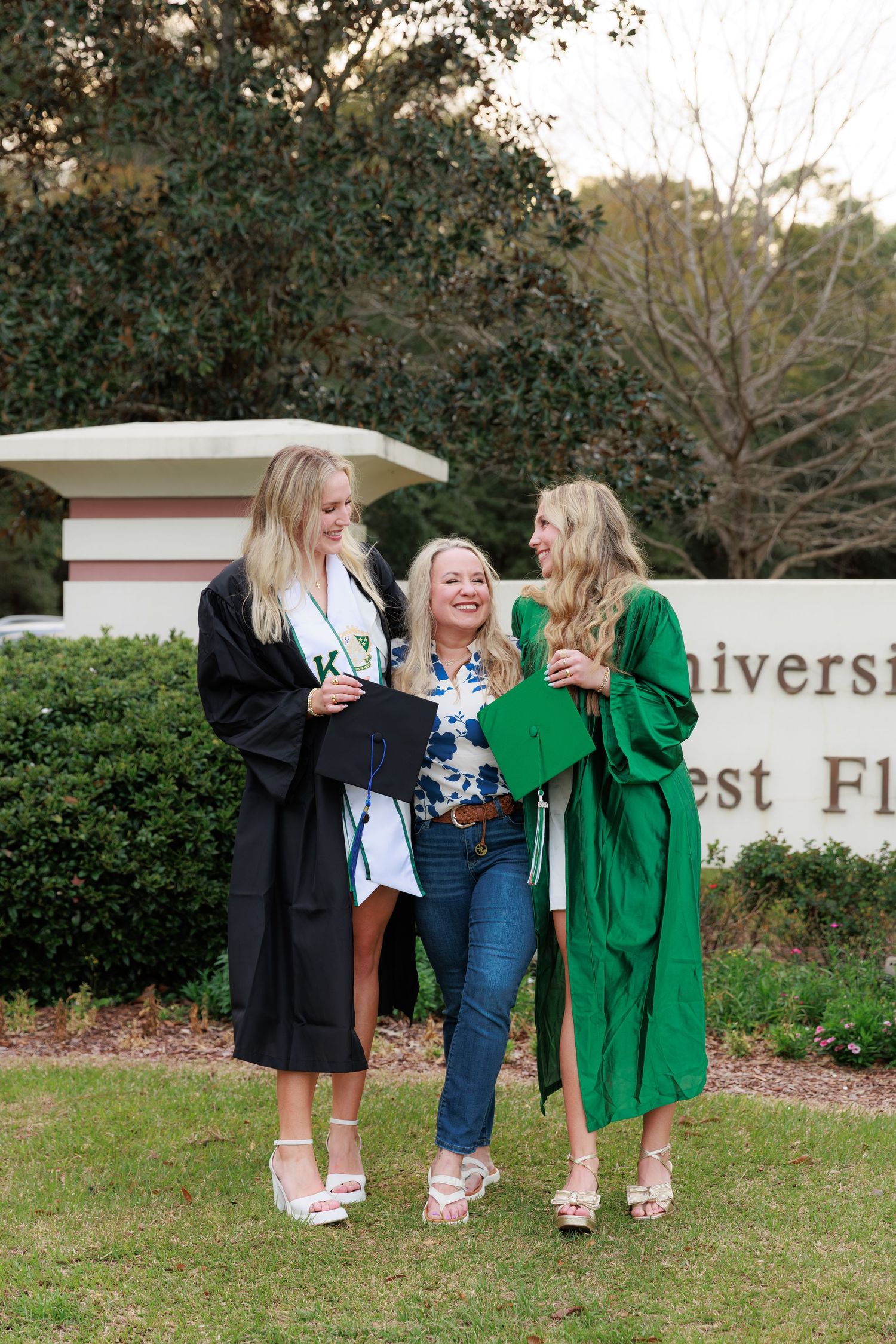Two female graduates in black and green robes celebrate with a woman at University of West Florida.