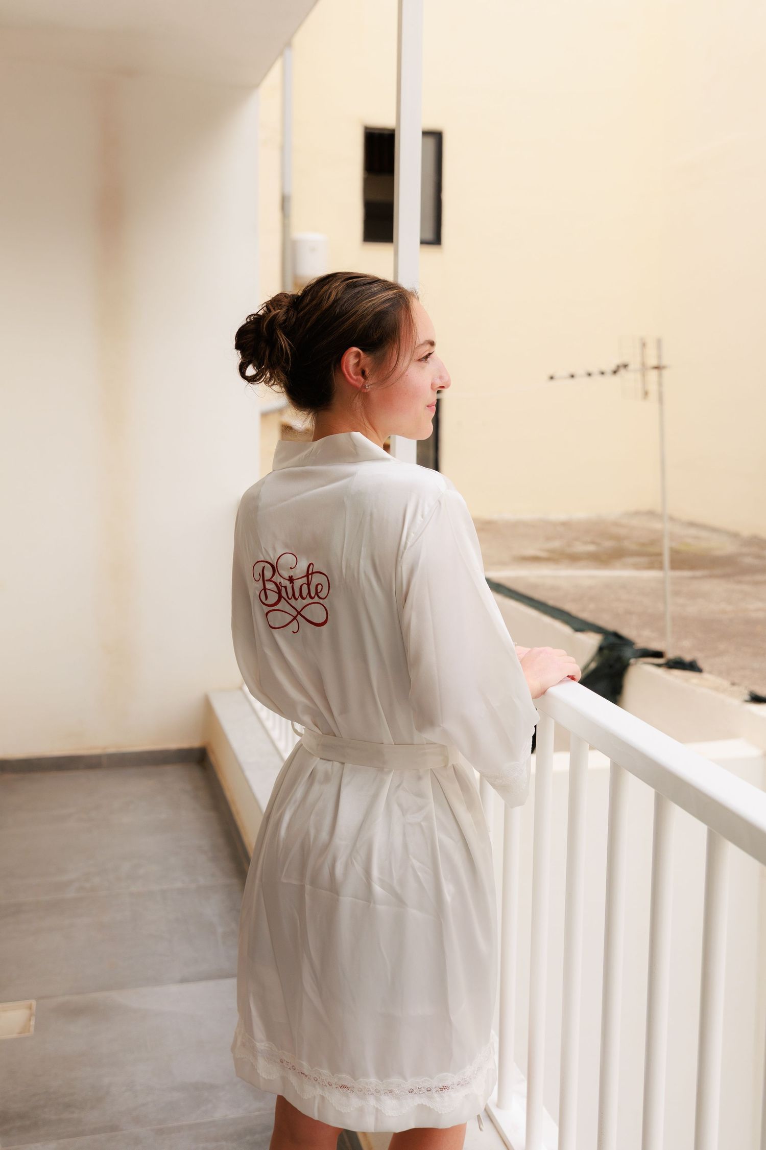 Woman in white robe standing on balcony looking at view during morning preparations.