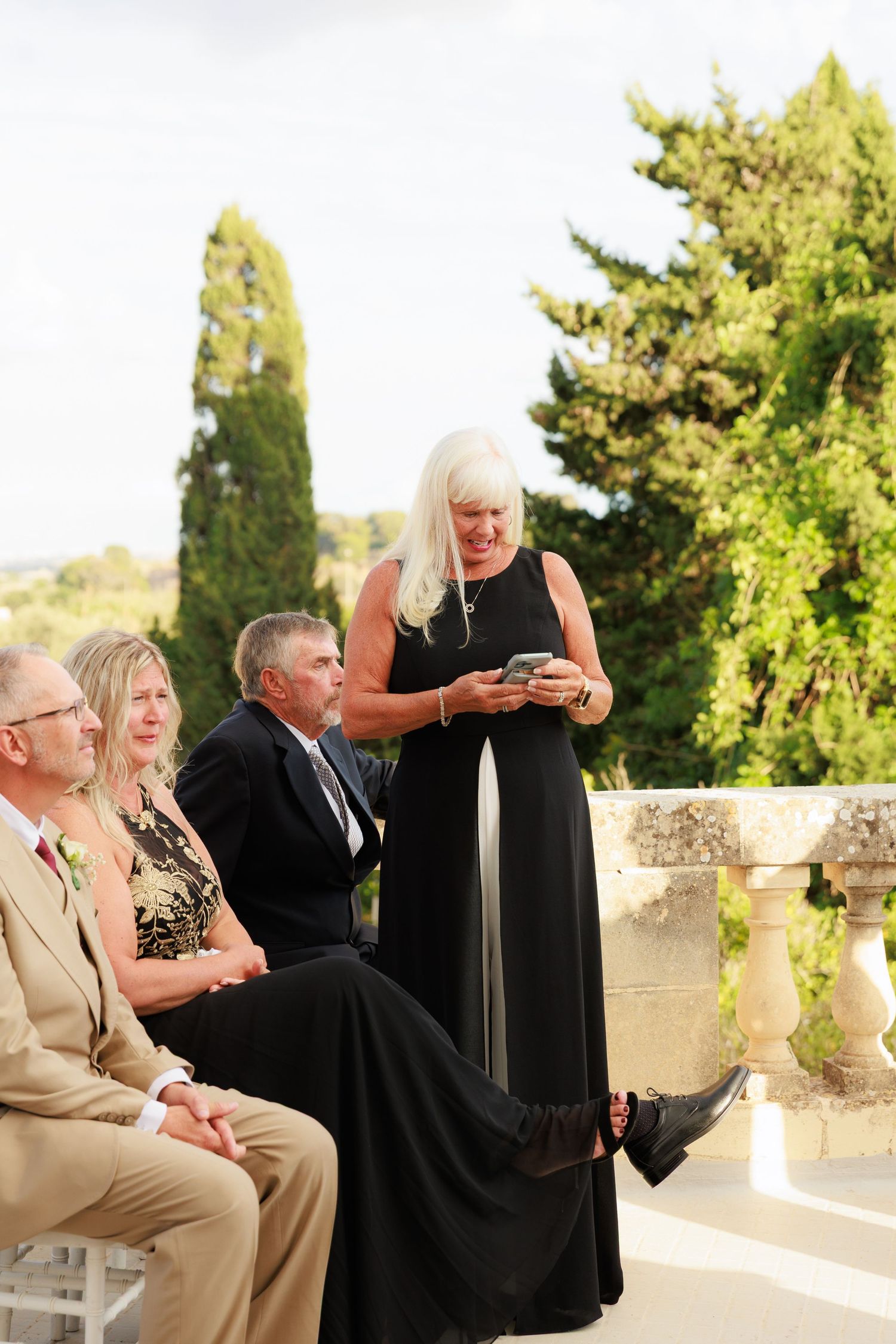 A group of people in formal attire gather on a sunny terrace with white stone balustrades and green trees in background.
