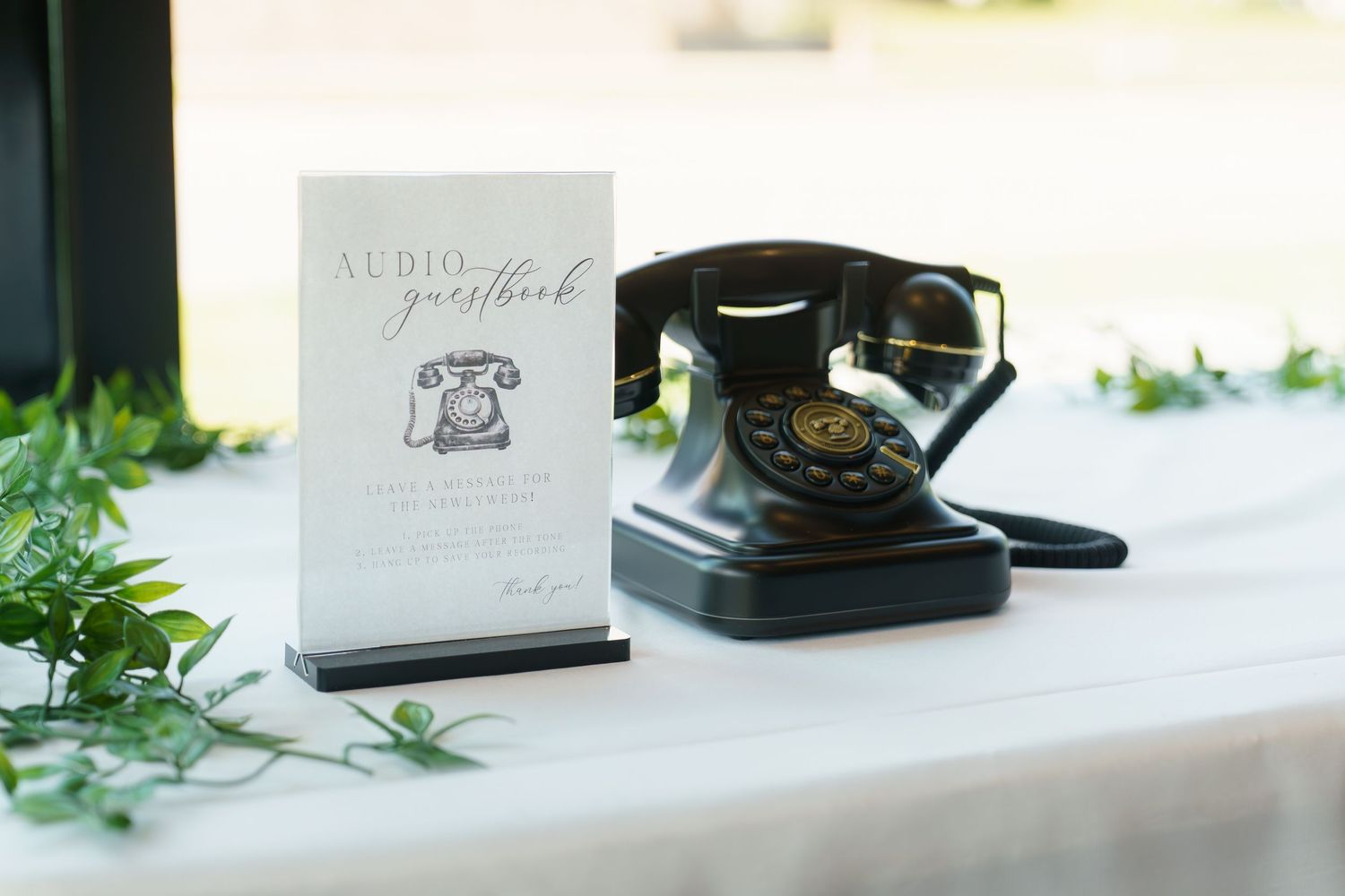 A vintage black rotary telephone sits on a white surface next to a menu card and decorative greenery.