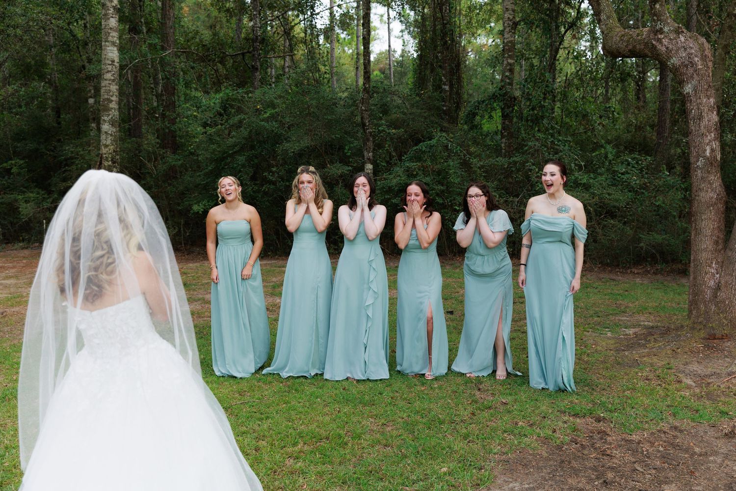 Wedding party wearing light blue bridesmaid dresses poses together in a wooded outdoor setting.