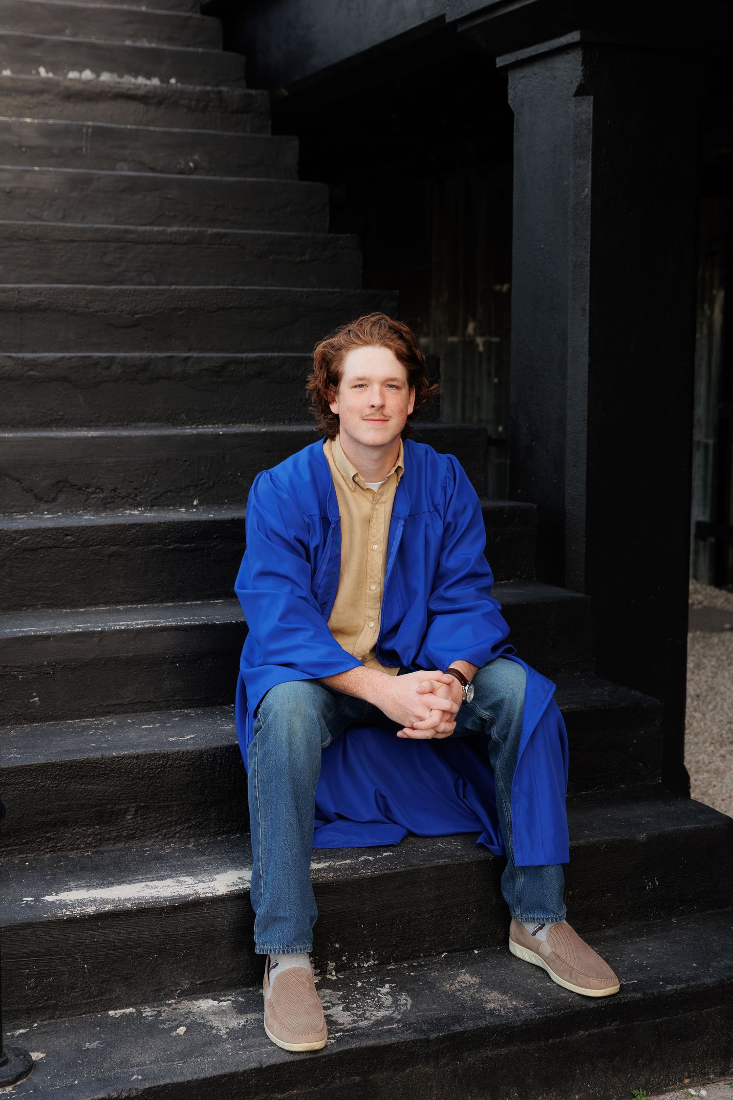 Young man in blue graduation gown sitting on dark outdoor steps.