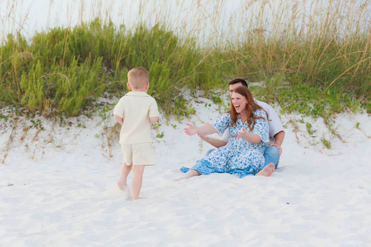 A woman in a blue floral dress sits on white sand beach dunes as a toddler boy walks toward her.