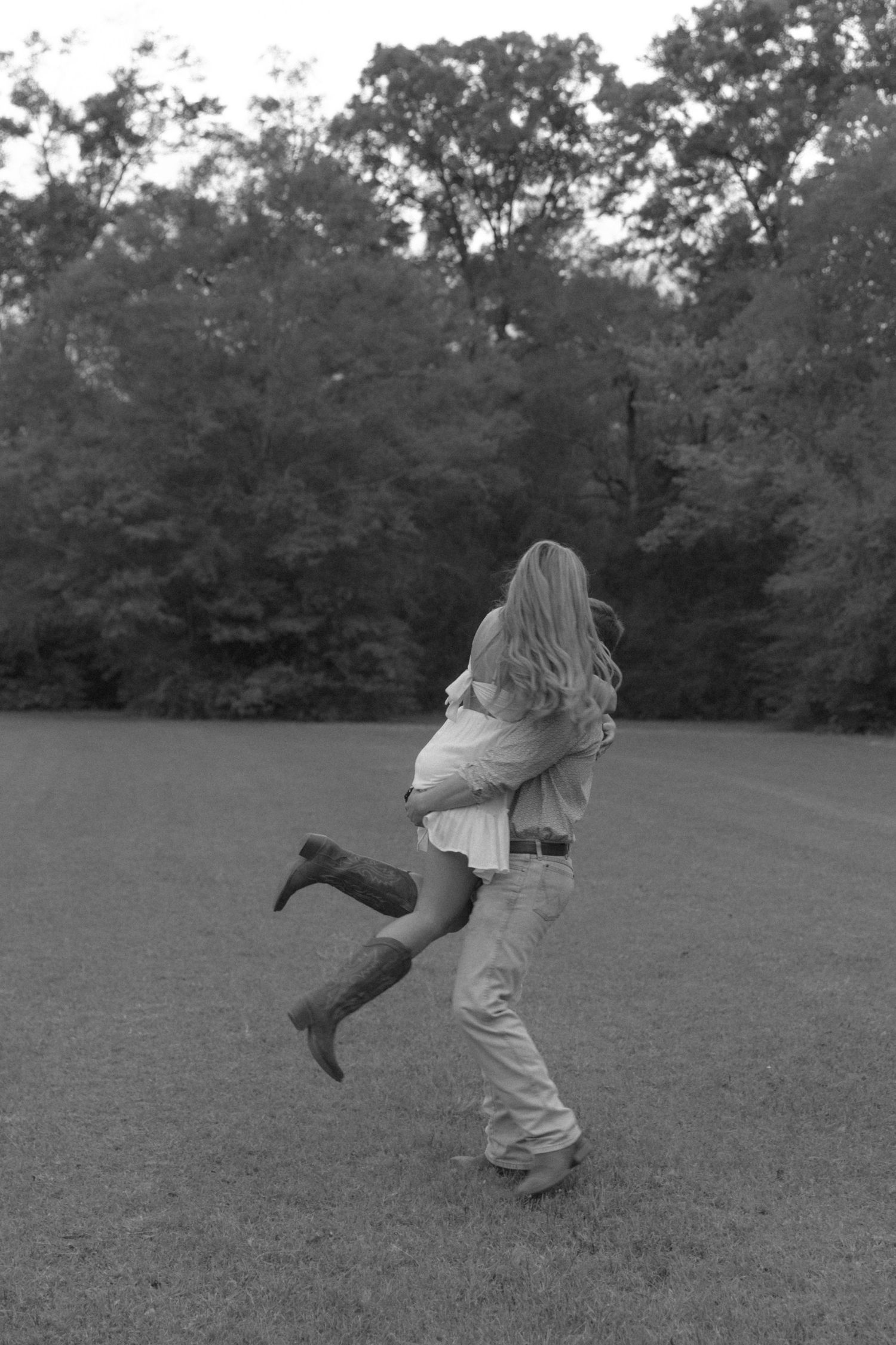 Black and white photo of a couple embracing in a grassy field, woman in cowboy boots lifted off ground.