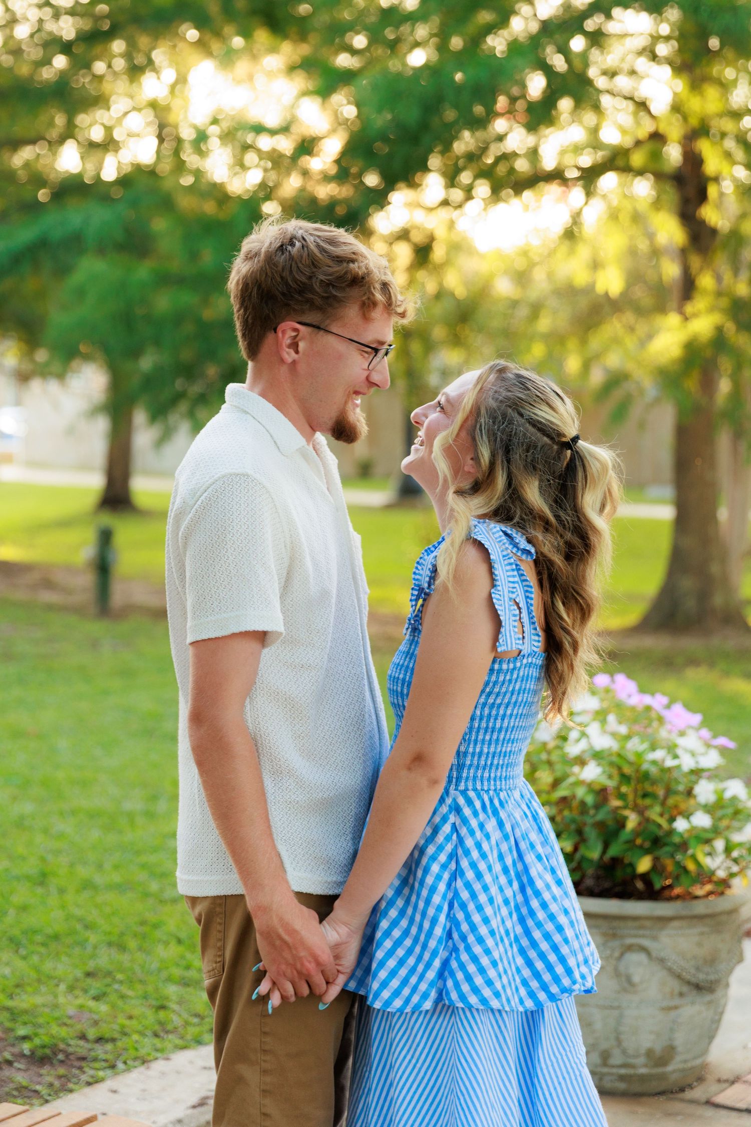 A happy young couple holds hands and laughs together outdoors in a lush green park, wearing casual summer outfits.